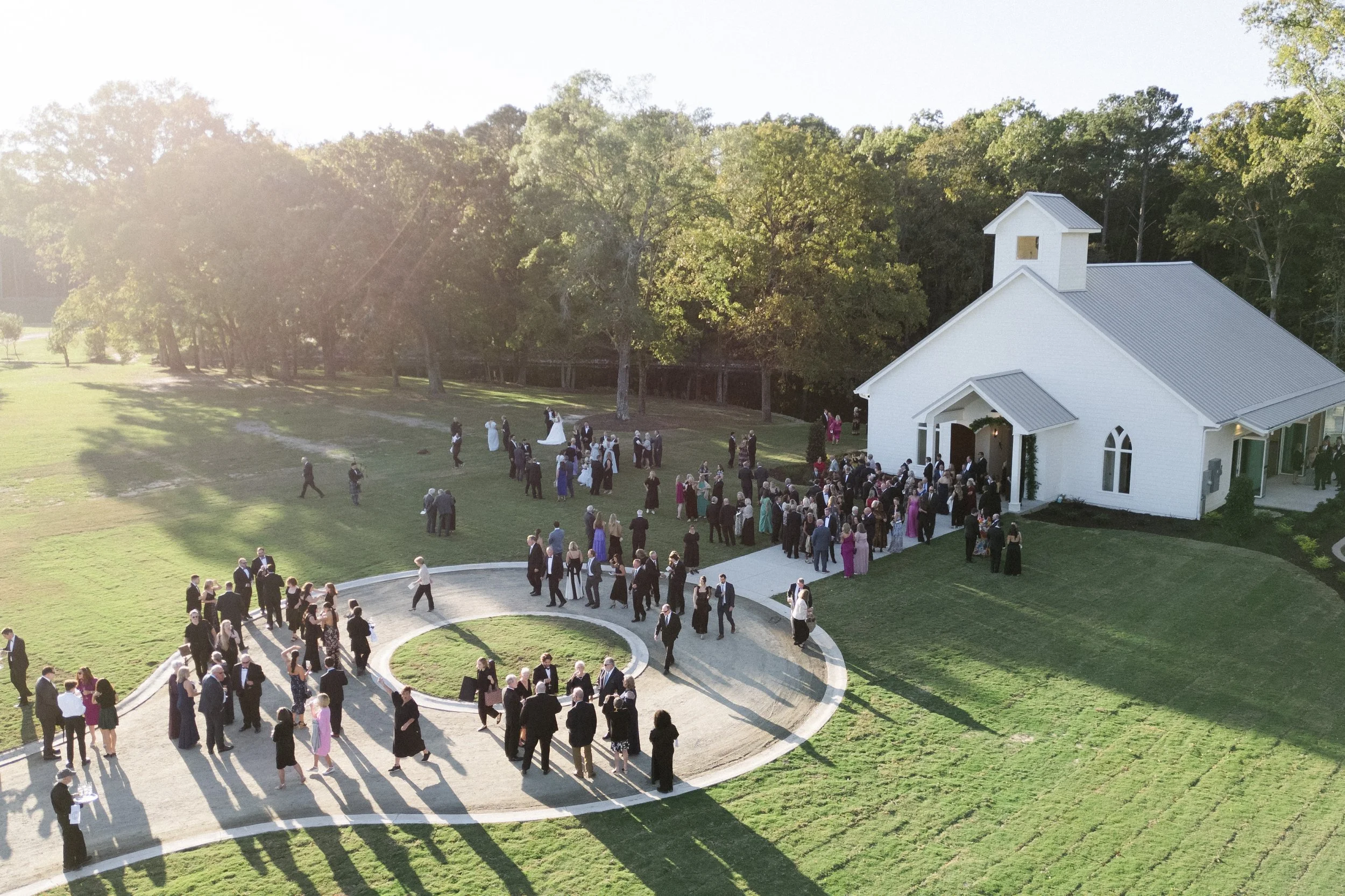 A large group of people gathered outside a white church with a small steeple, surrounded by green grass and trees, on a sunny day.