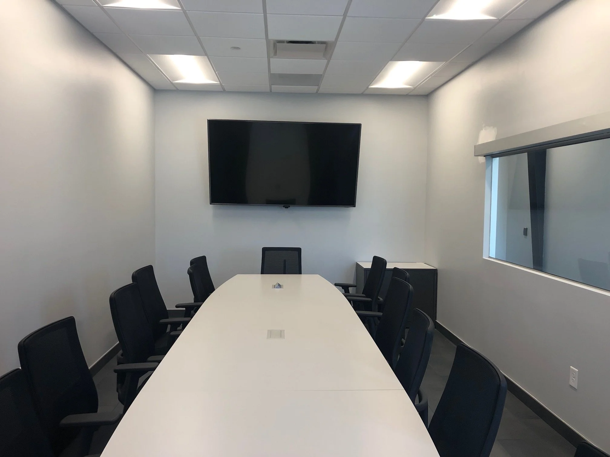 Empty conference room with a long white table, black chairs, a wall-mounted flat-screen TV, a window with blinds, white walls, and ceiling lights.