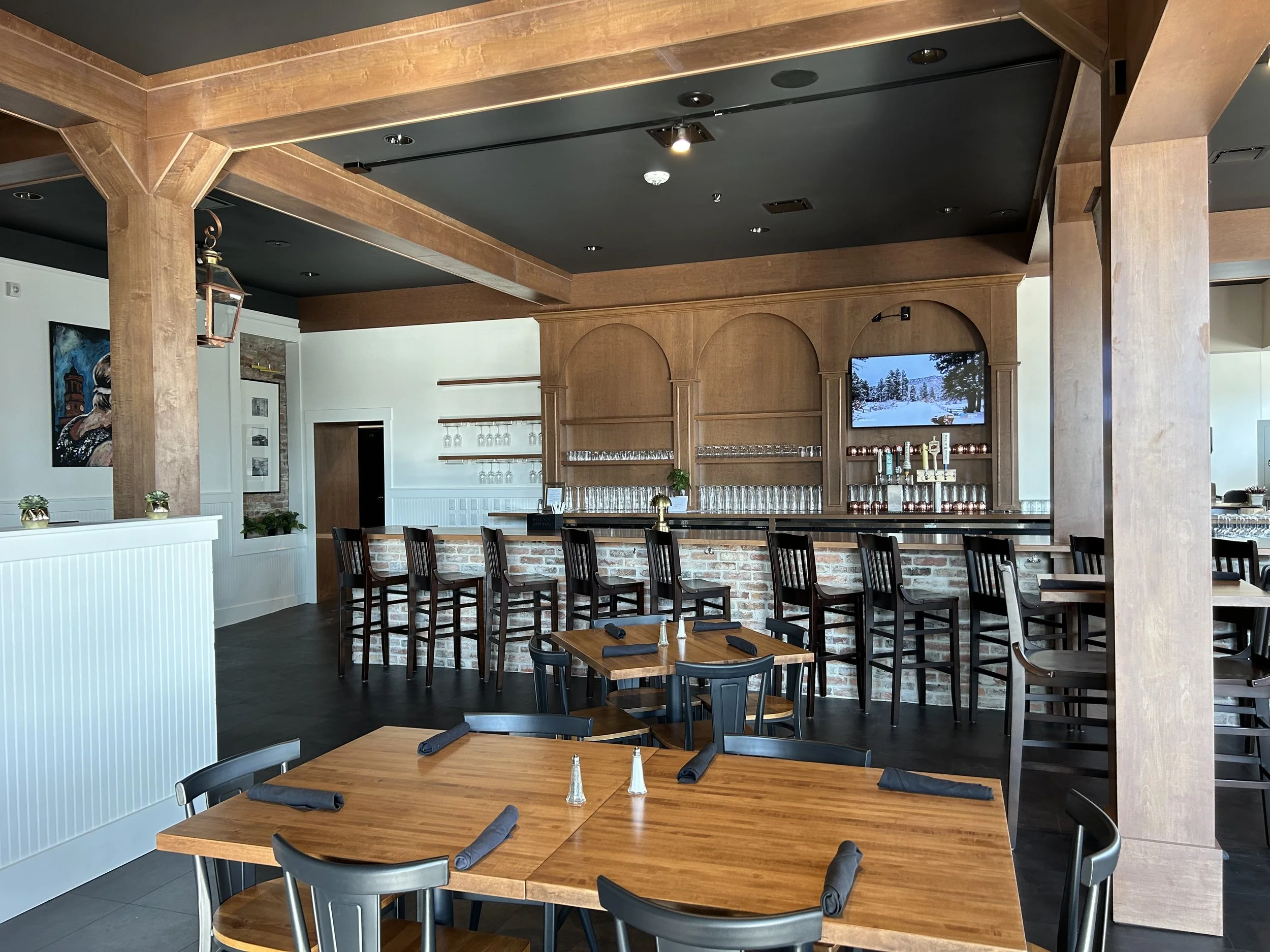 Interior view of a restaurant with wooden tables and chairs, a bar area with glasses, a large screen displaying snowy landscape, and artwork on the walls.