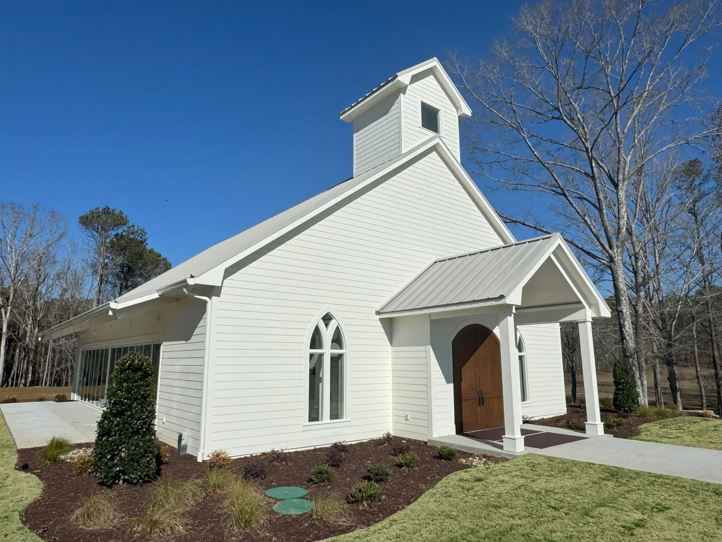 A white church with a wooden door, arched windows, and a steeple, surrounded by a landscaped lawn with bushes and trees under a clear blue sky.