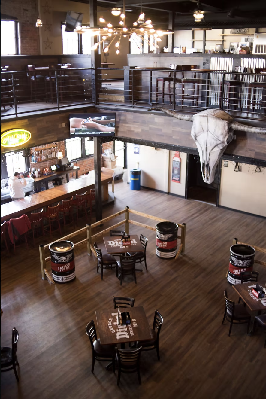 Interior of a rustic bar or restaurant with wooden floors, a bar counter with chairs, a large animal skull mounted on the wall, and a television mounted above the bar. There is a chandelier hanging from the ceiling, and tables with chairs are arrange