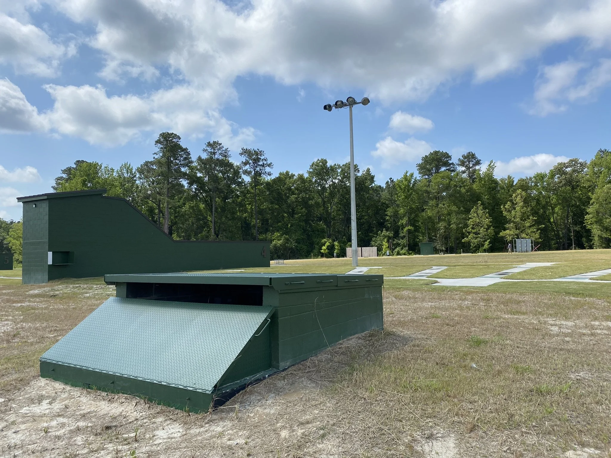 Outdoor athletic field with green equipment and a tall light pole, surrounded by trees and a partly cloudy sky.