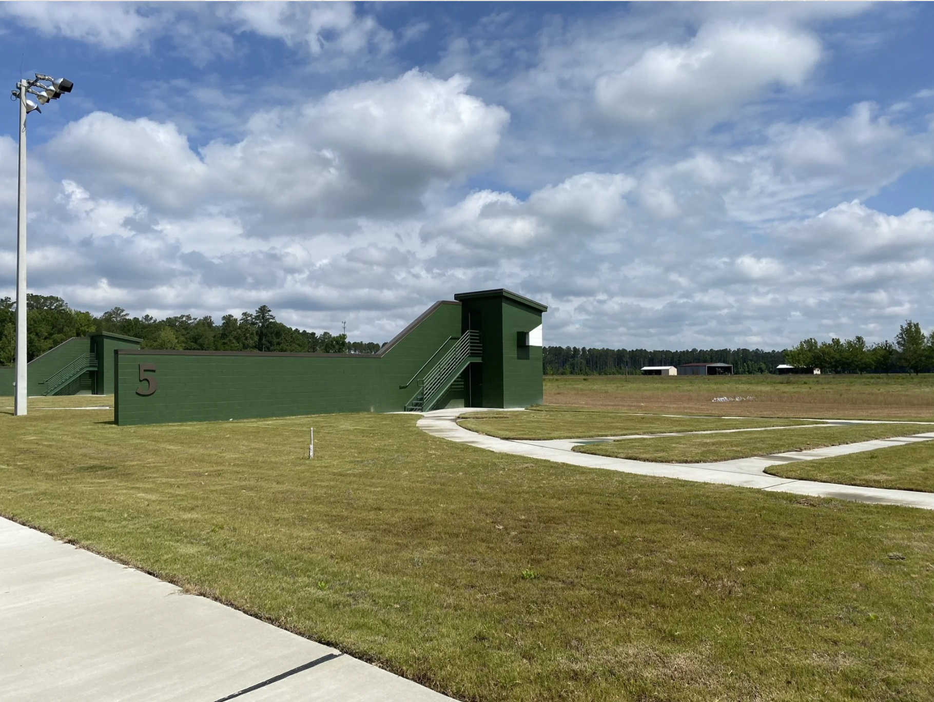 A green sports training facility with a staircase, a tall pole with floodlights, and a grassy field under a partly cloudy sky.