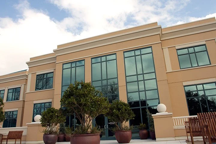 A modern beige-colored building with large glass windows and doors, potted plants with trees in front, and outdoor benches, under a partly cloudy sky.