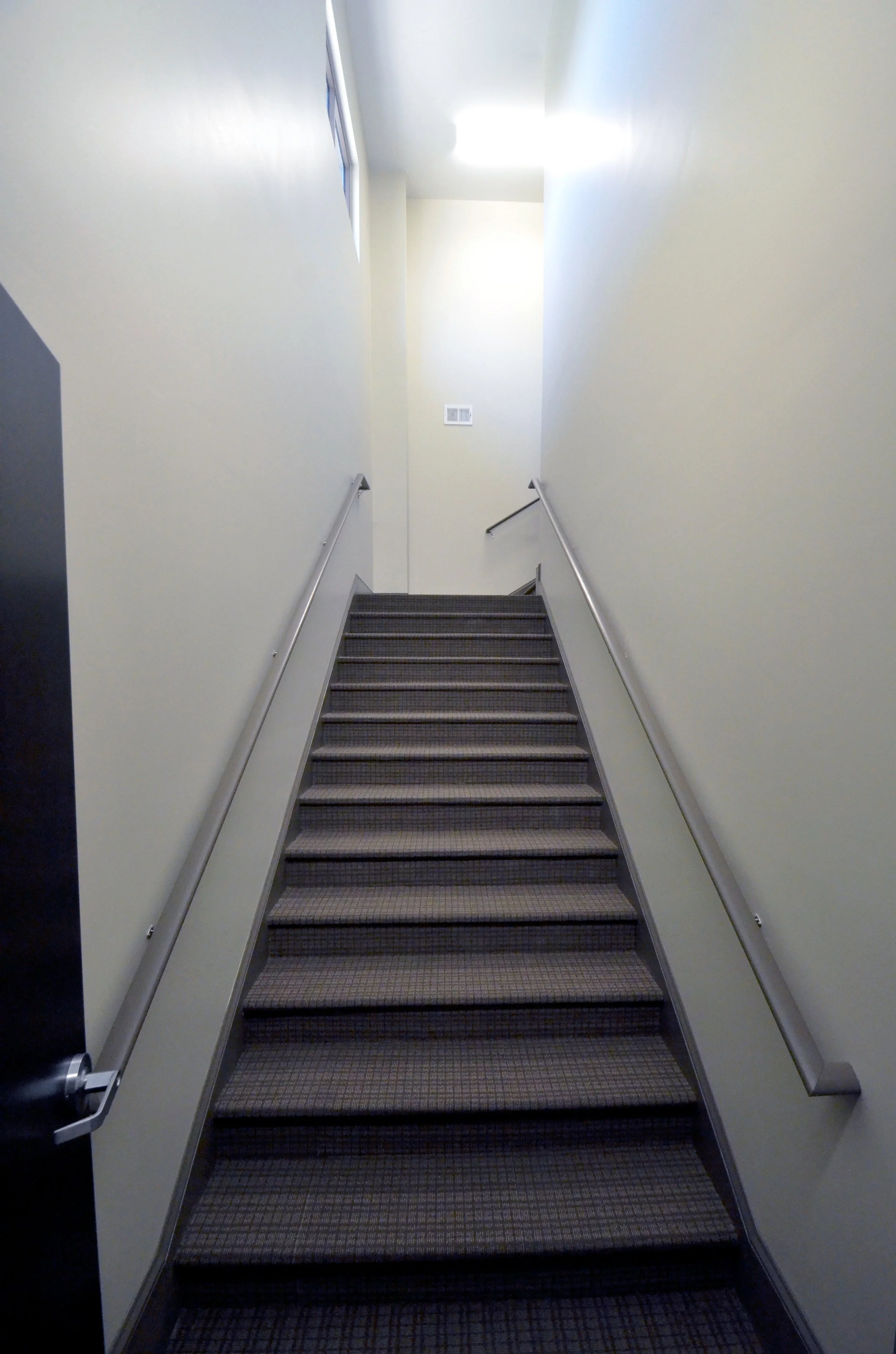 Indoor staircase with dark carpeting, beige walls, metal handrails on both sides, leading upwards towards a well-lit area with a wall vent visible on the right wall at the top of the stairs.
