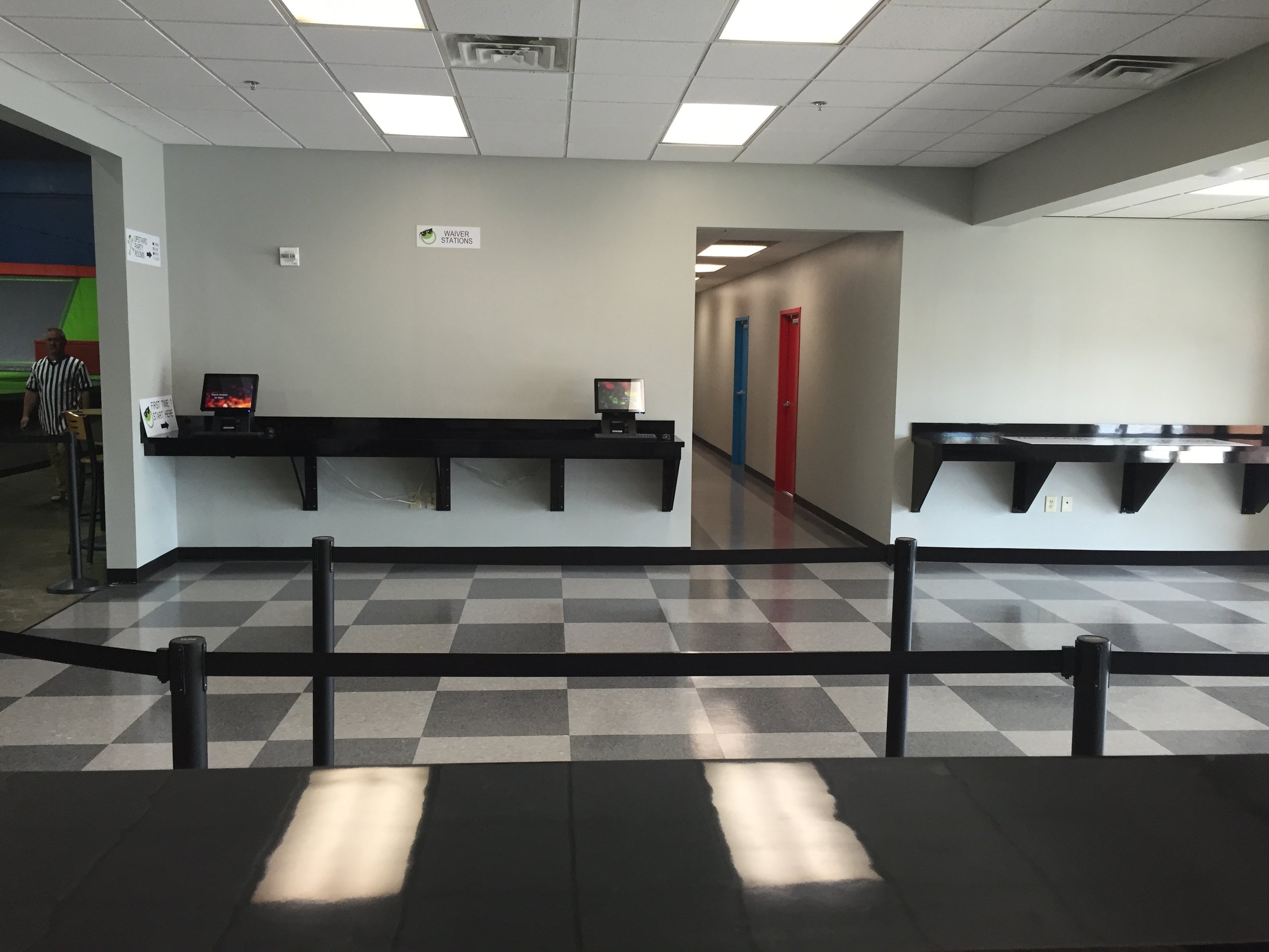 Empty check-in area with two touchscreen kiosks on a black counter and a sign for waiver stations on the beige wall behind. Visible entrance with black stanchions and black-and-gray checkered tile floor.
