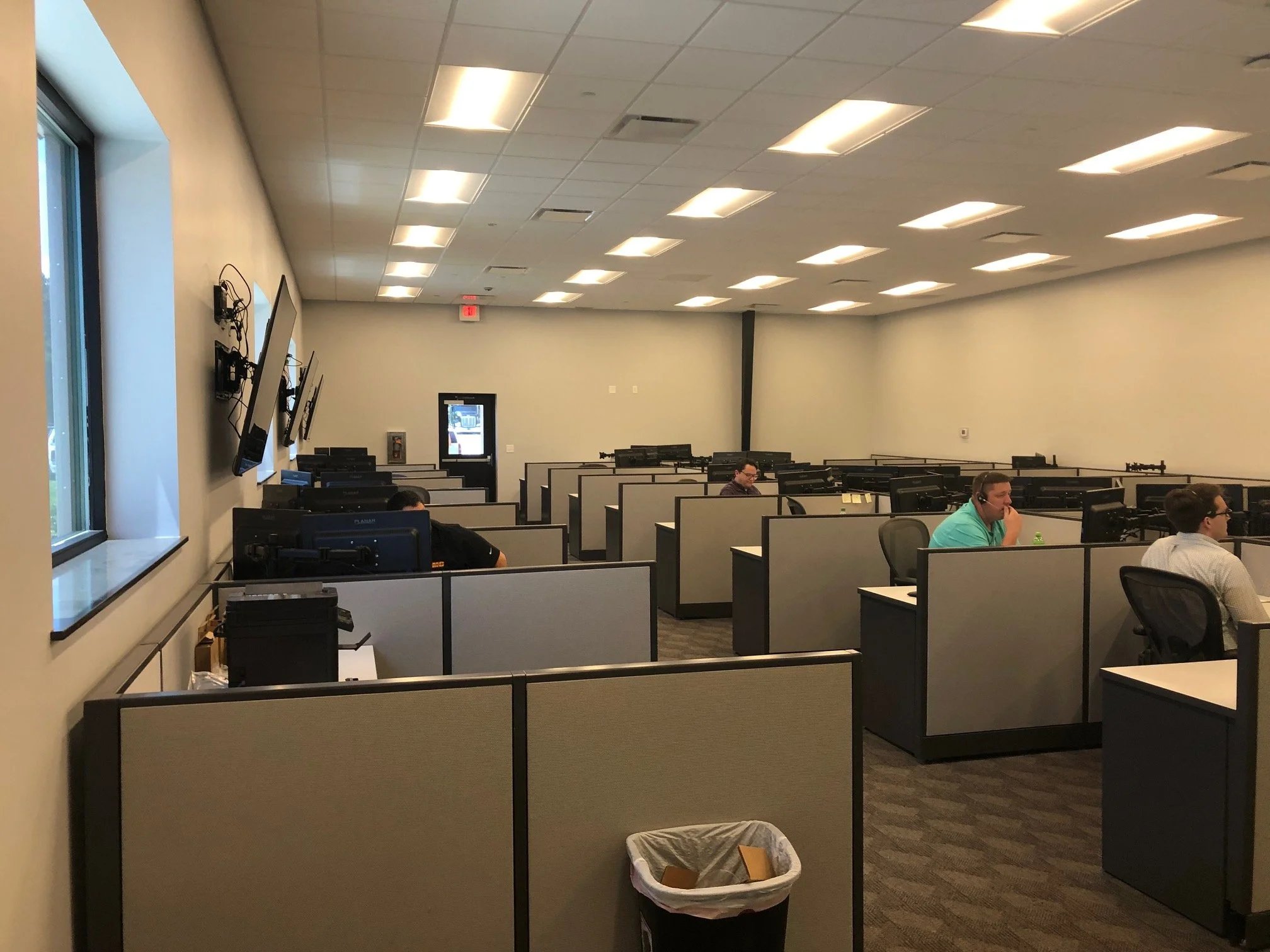 A computer lab with rows of cubicles, each with a person working on a computer. There are multiple television screens mounted on the beige wall, and a window on the left. Fluorescent ceiling lights illuminate the room.