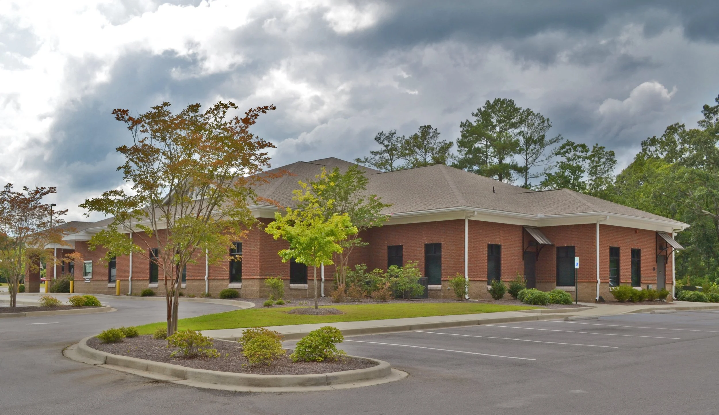 A brick building with a gray shingle roof, surrounded by trees and small shrubs, under a cloudy sky. There is a parking lot in the foreground with parking spaces and curbs.