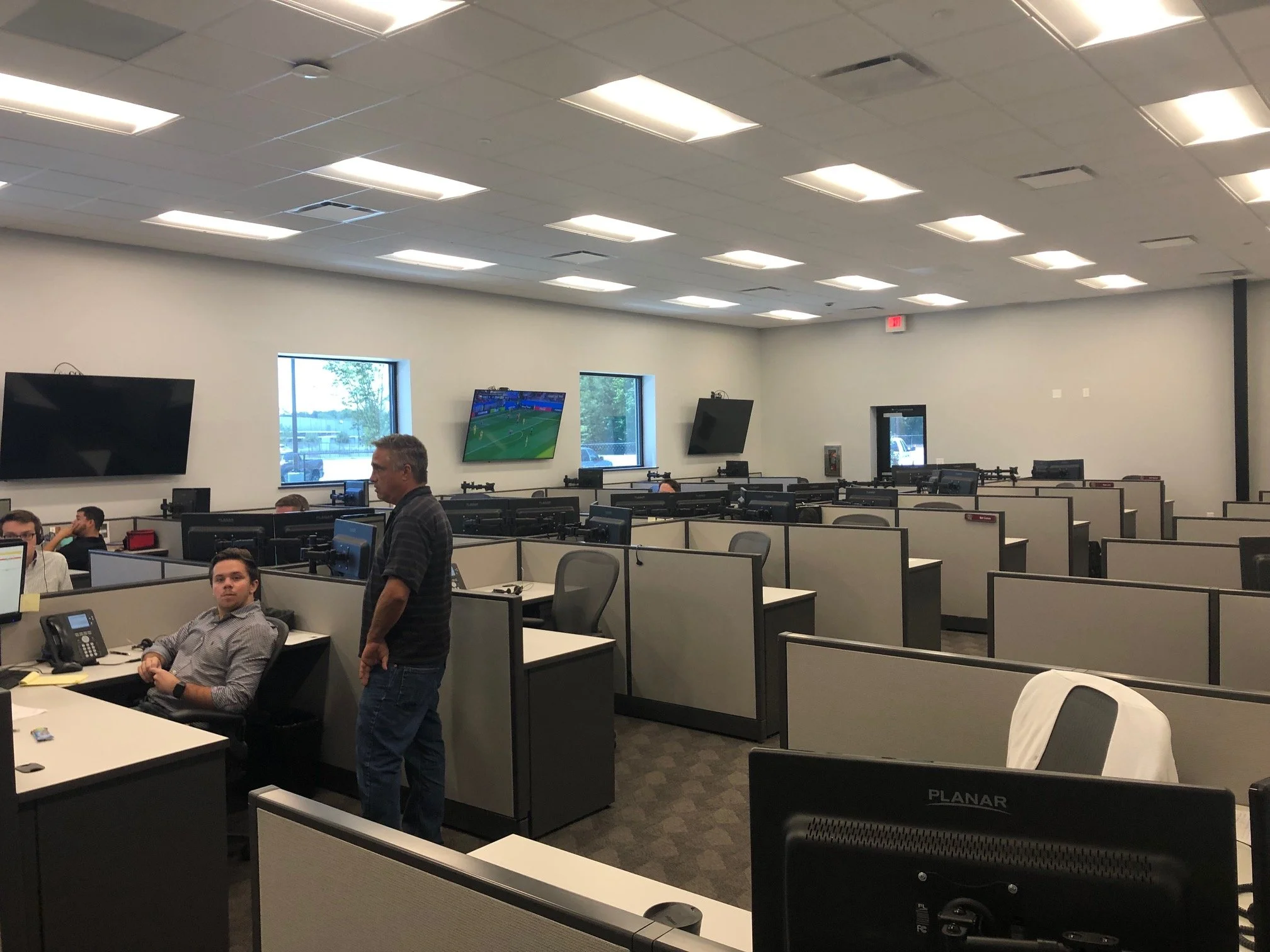An office with multiple rows of cubicles, several people working at desks, two large wall-mounted monitors displaying a soccer game, and a man standing talking to another person. The ceiling has evenly spaced rectangular lights, and there are windows