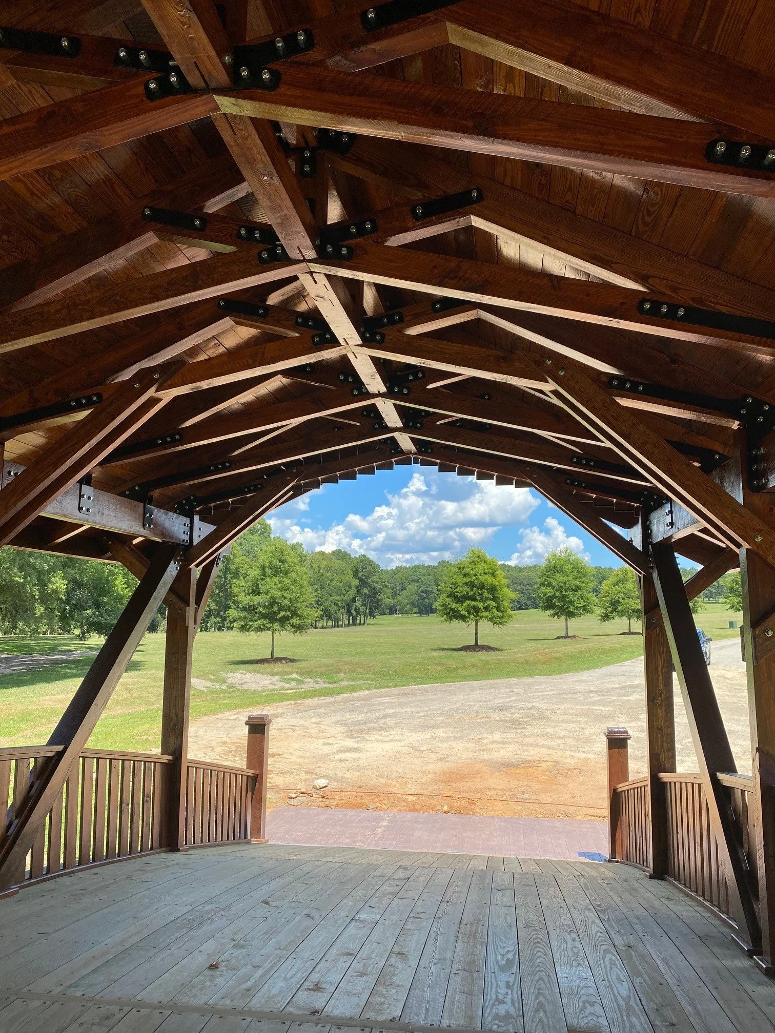 View from inside a wooden gazebo looking out onto a grassy field with trees and blue sky in the distance.