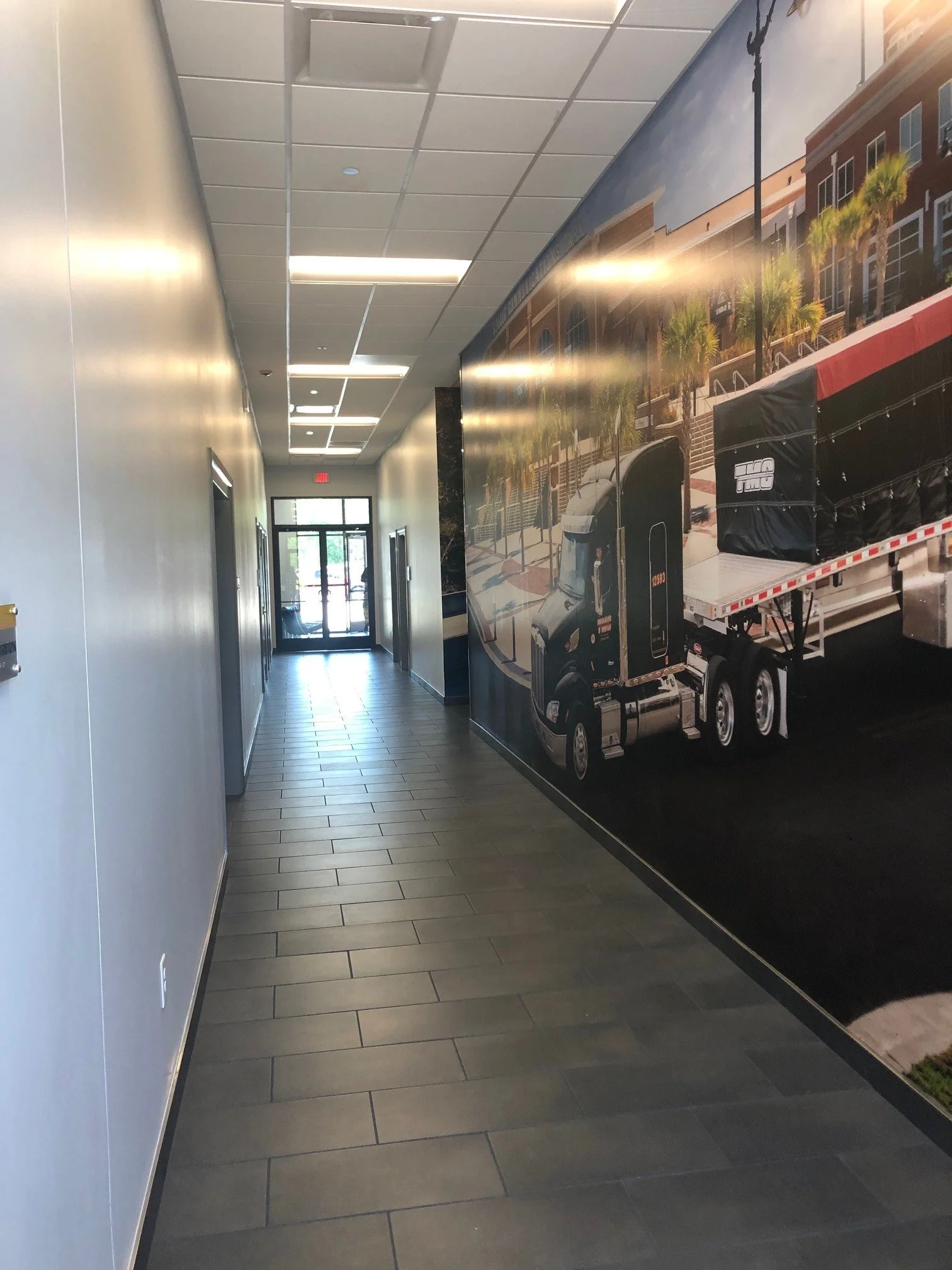 A corridor inside a building with tiled flooring, gray walls, and a ceiling with fluorescent lights. There is a large mural of a city street scene on one wall, featuring palm trees, buildings, and a truck.