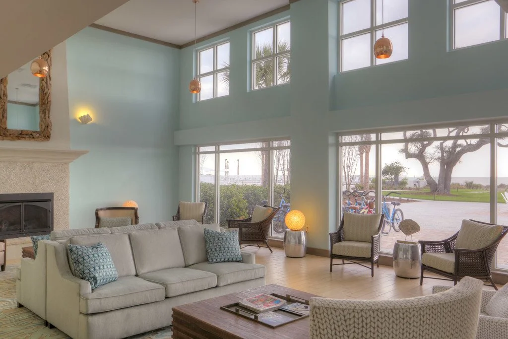 A spacious, modern living room with large floor-to-ceiling windows, light-colored furniture, a stone fireplace, and outdoor greenery visible through the windows.