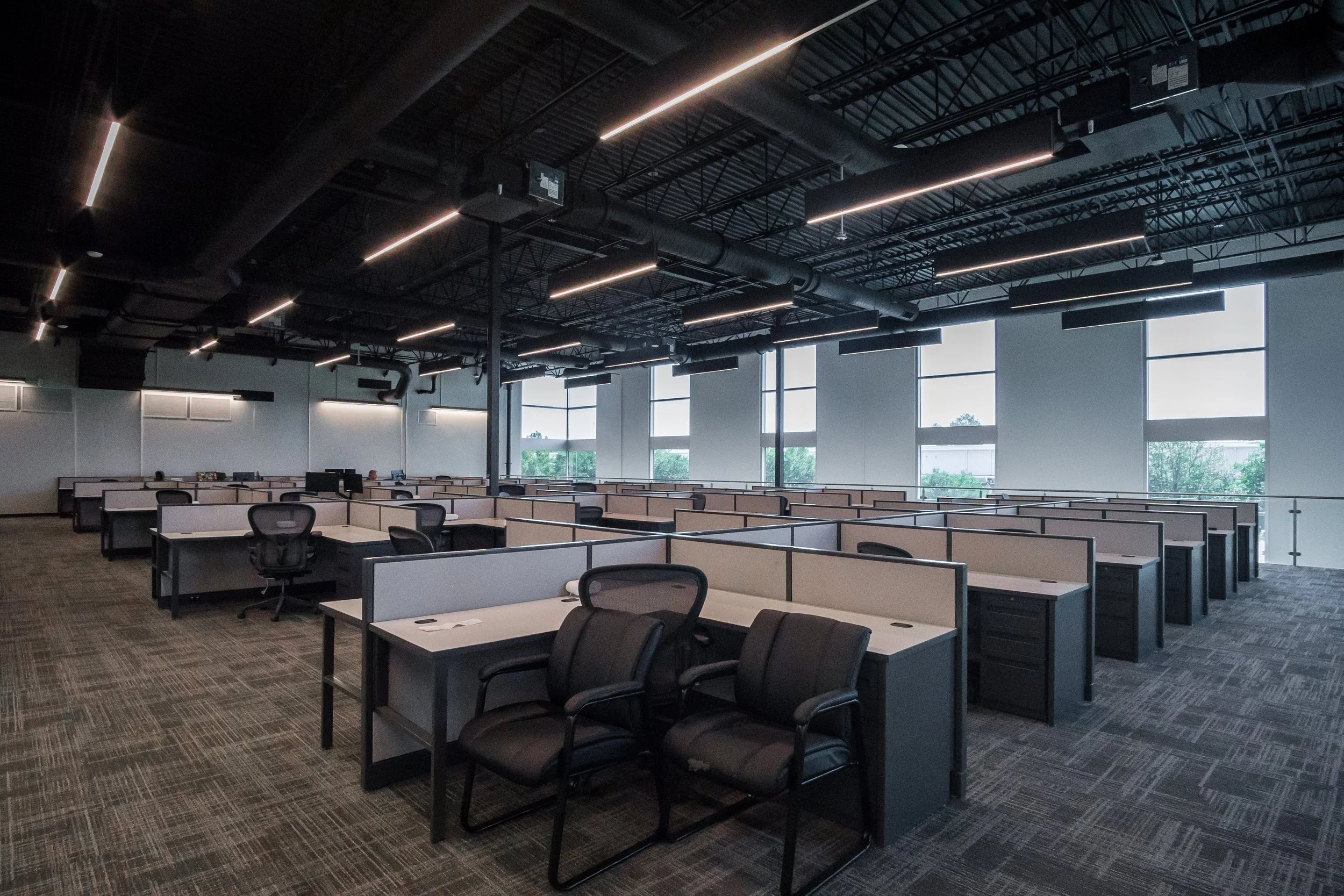 Large modern office space with multiple workstations, black office chairs, and large windows that let in natural light. The ceiling has exposed ductwork and long linear lighting fixtures.
