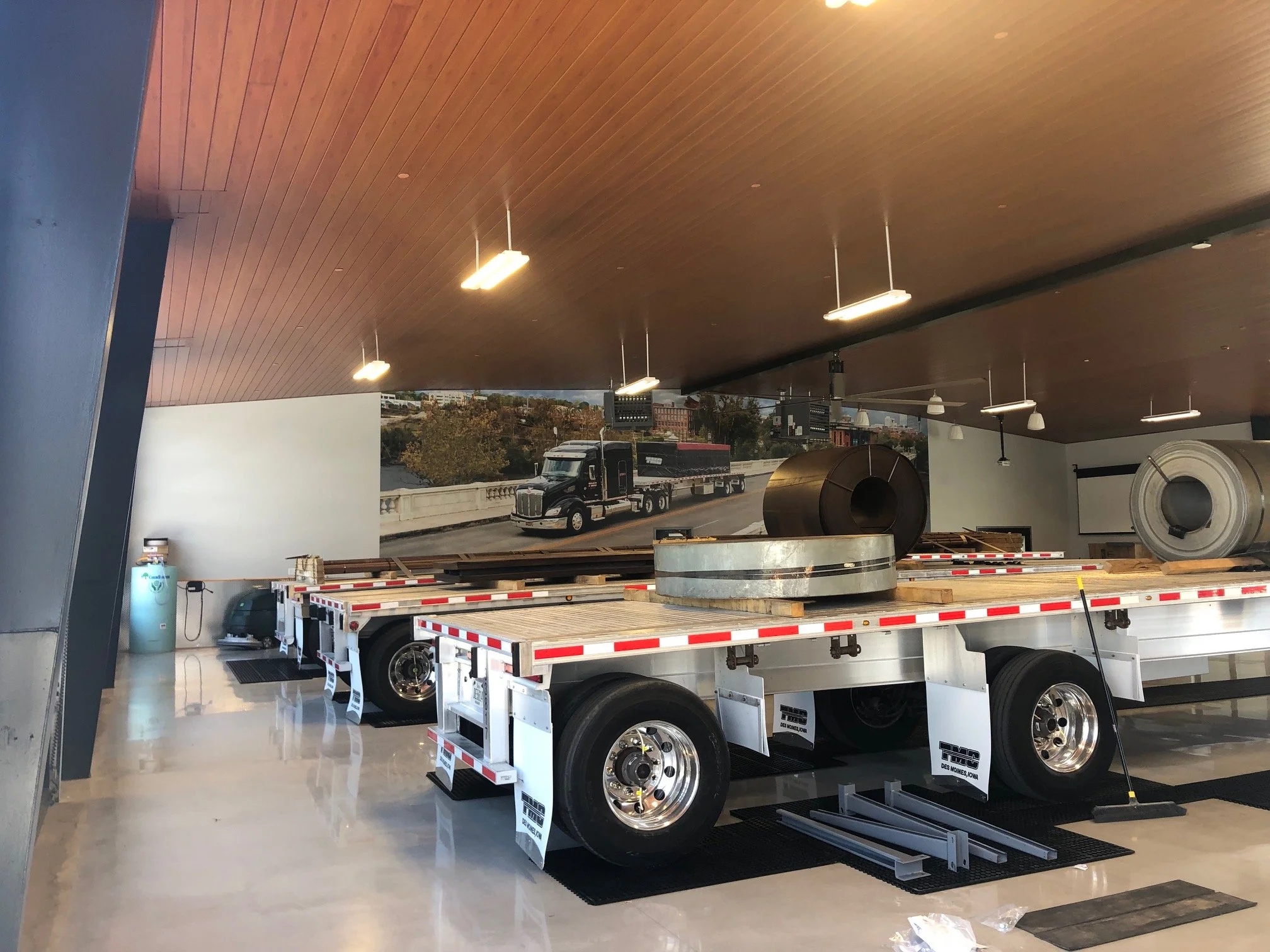 A flatbed trailer with metal wheels inside a showroom or warehouse. The background features large windows showing a highway with trucks and buildings outside. The trailer is unhooked and placed on a shiny, reflective floor.