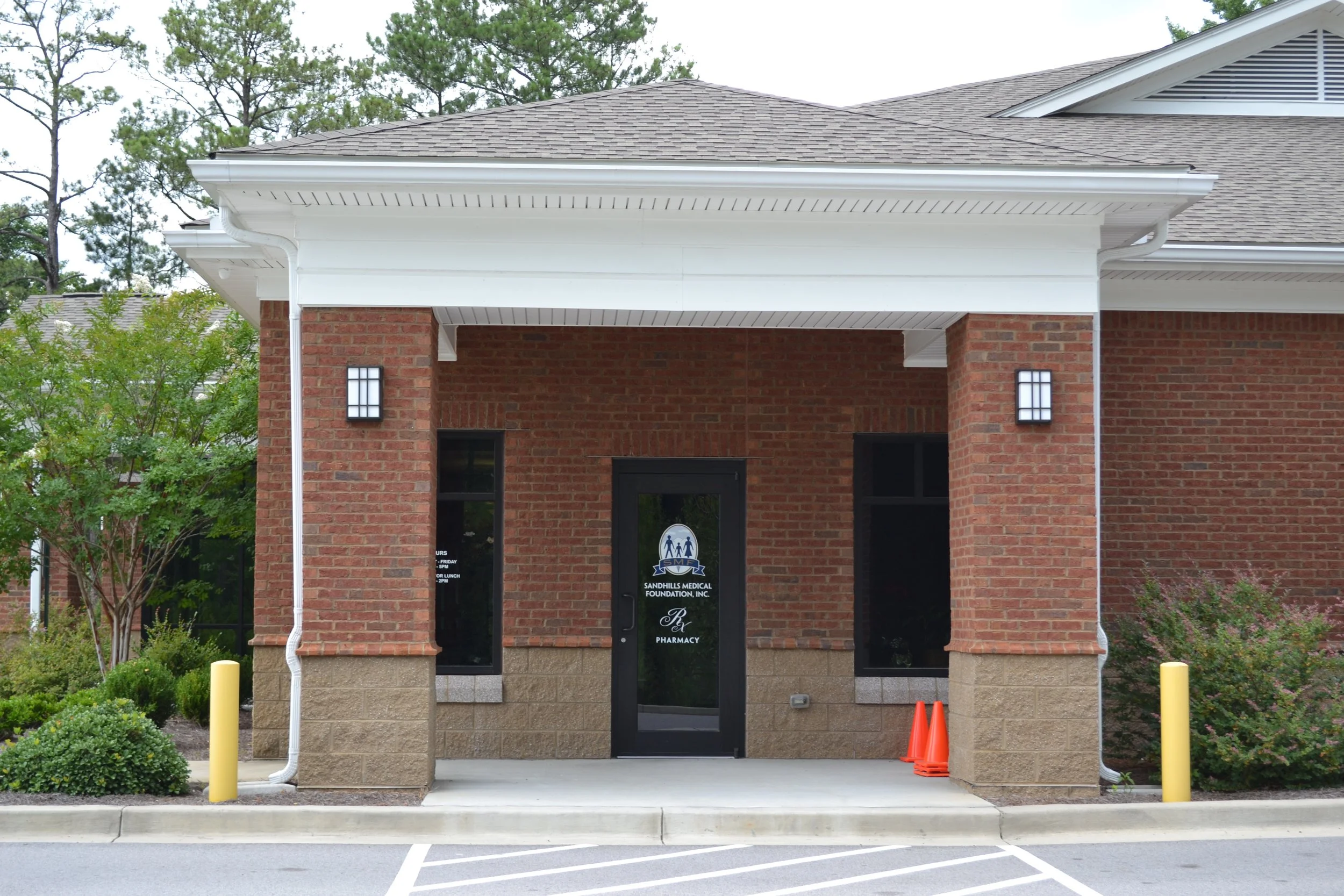 Front of a brick building with a sign indicating a pharmacy and medical foundation, flanked by yellow posts and orange cones, with greenery and trees in the background.