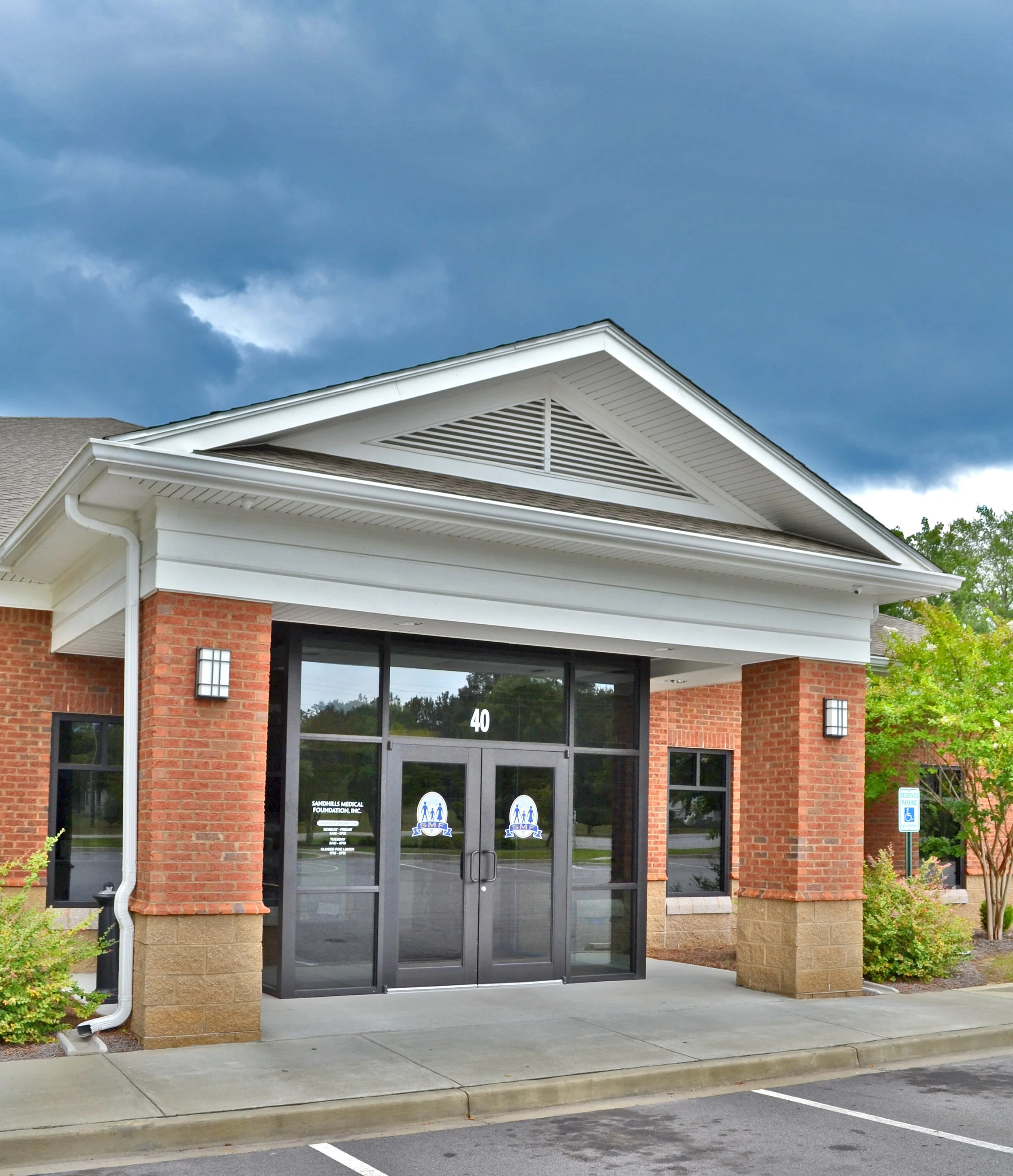 Front entrance of a building with double glass doors, brick and stone exterior, and a sign on the door for Sands Medical Foundation, with a handicap parking sign on the right and trees in the background.
