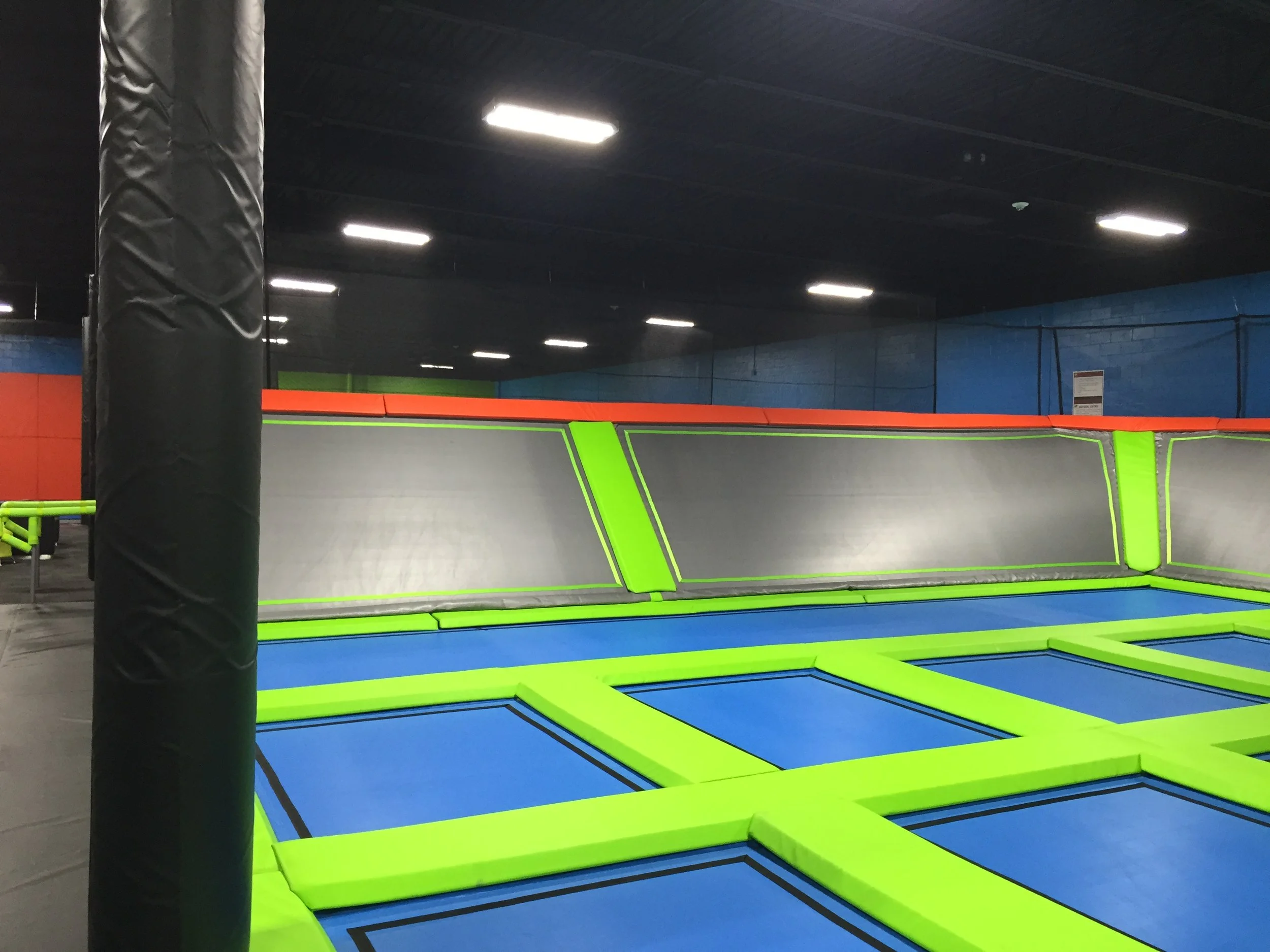 Indoor trampoline park with a large area of interconnected blue and green trampolines, padded safety zones, and black vertical padding around support poles.