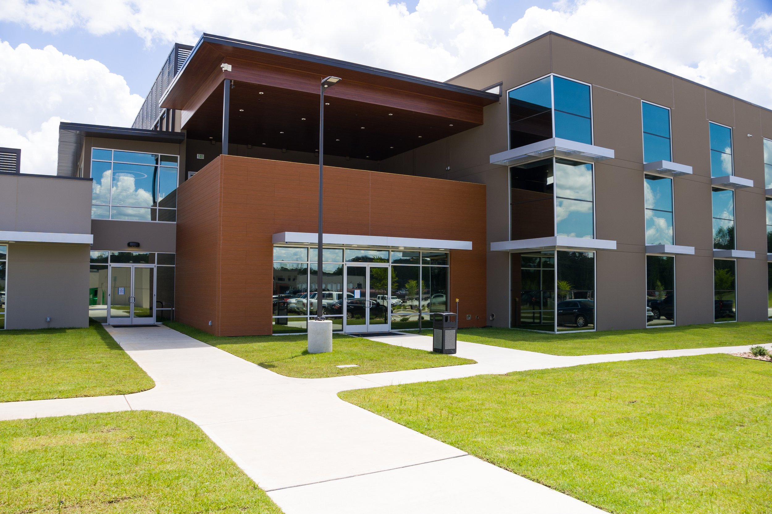 Modern multi-story office building with large glass windows, a brown and gray exterior, and a green lawn in front under partly cloudy sky.
