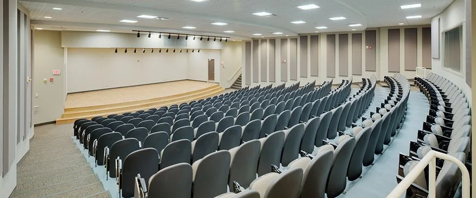 Empty auditorium with rows of black and white chairs facing a stage.