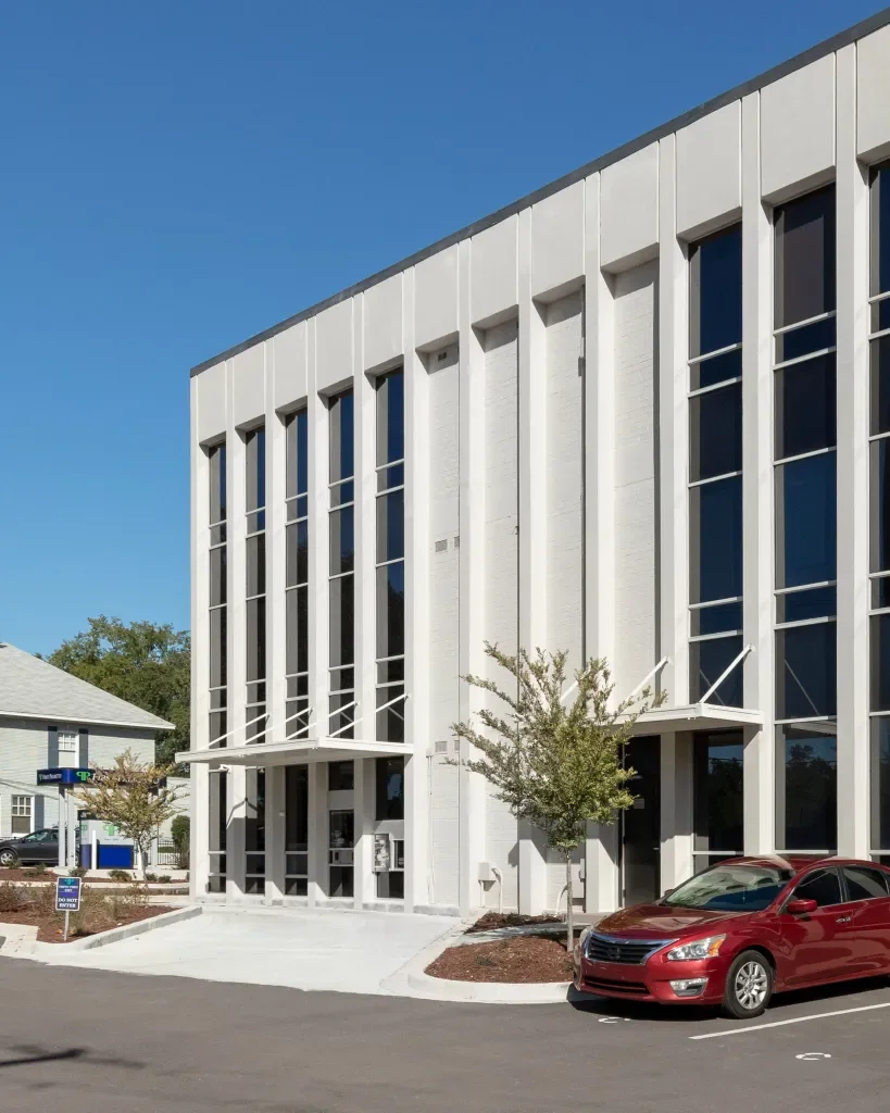 Modern white building with tall vertical windows and small trees in front, parked red car, clear blue sky.