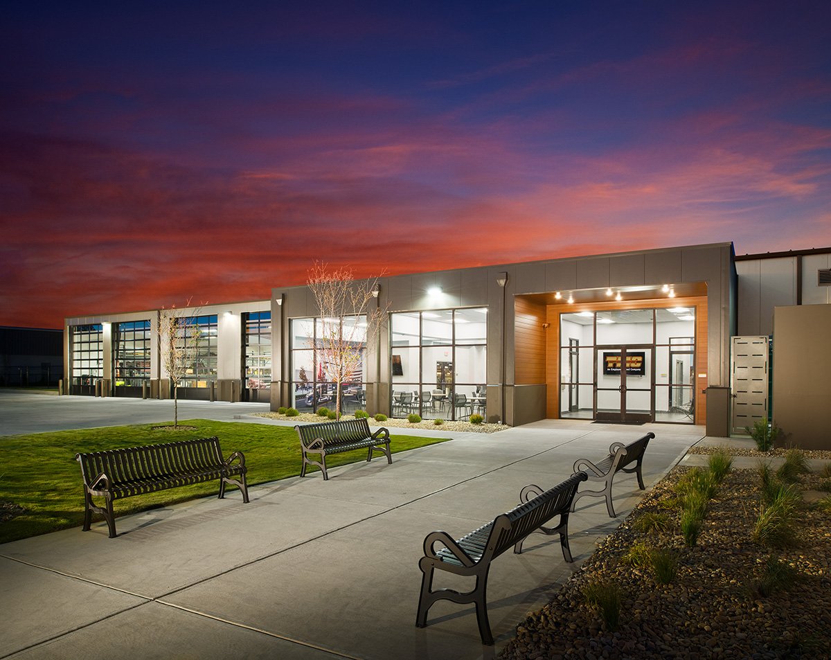 Modern building in the evening with large glass windows, seating benches, trees, and a colorful sunset sky.