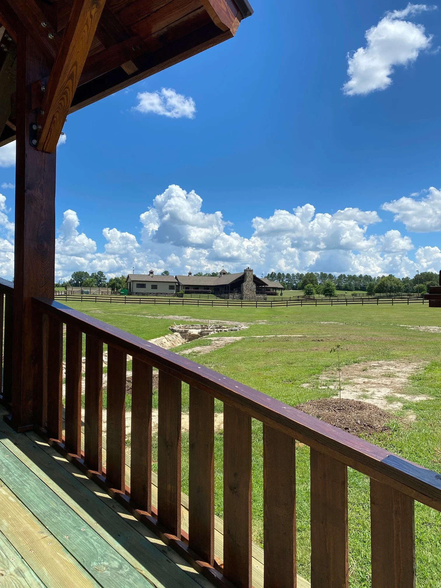 View from a wooden porch or balcony looking out over a large grassy field with a fenced area, a house with a chimney, and a stone structure in the distance; partly cloudy sky with bright blue background.