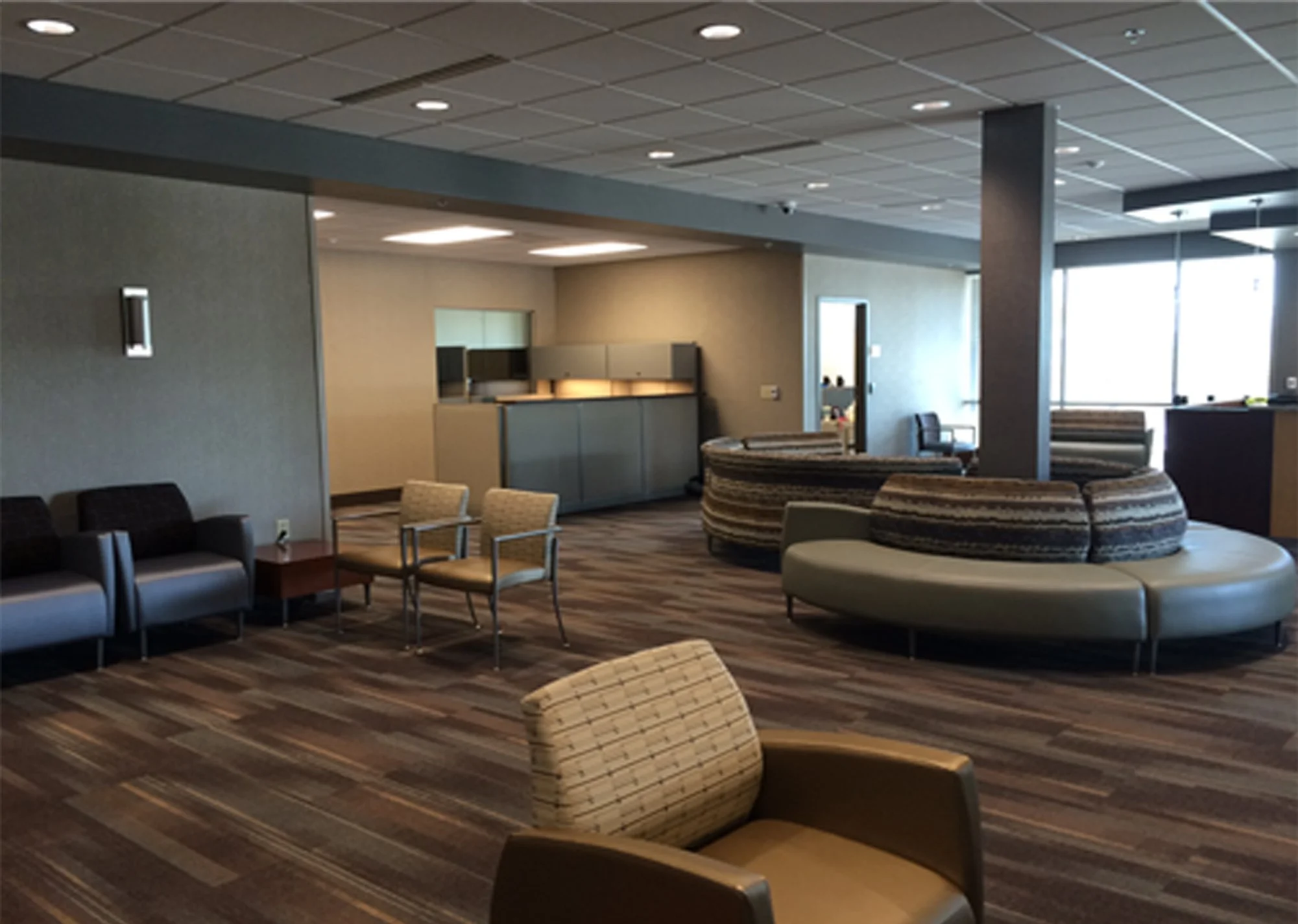 Interior of a modern lounge or waiting area with chairs and sofas, a carpeted floor, and large windows allowing natural light, with a reception area in the background.