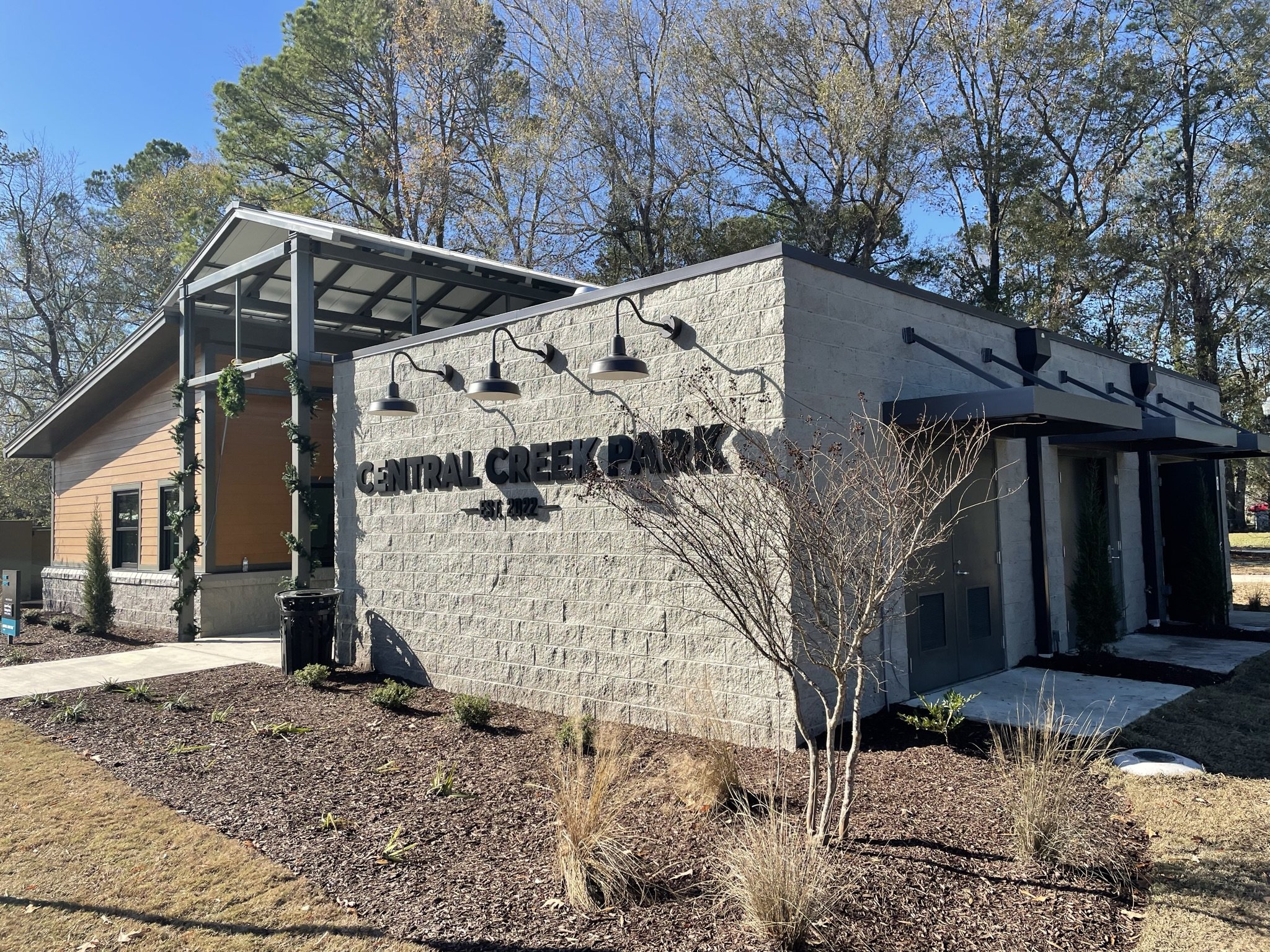 A modern building with a sign that reads 'Central Creek Park Est. 2022'. The building has a light-colored brick exterior, black metal railings, and hanging lights. Surrounding the building are trees, some with leaves and others without, and a landsca