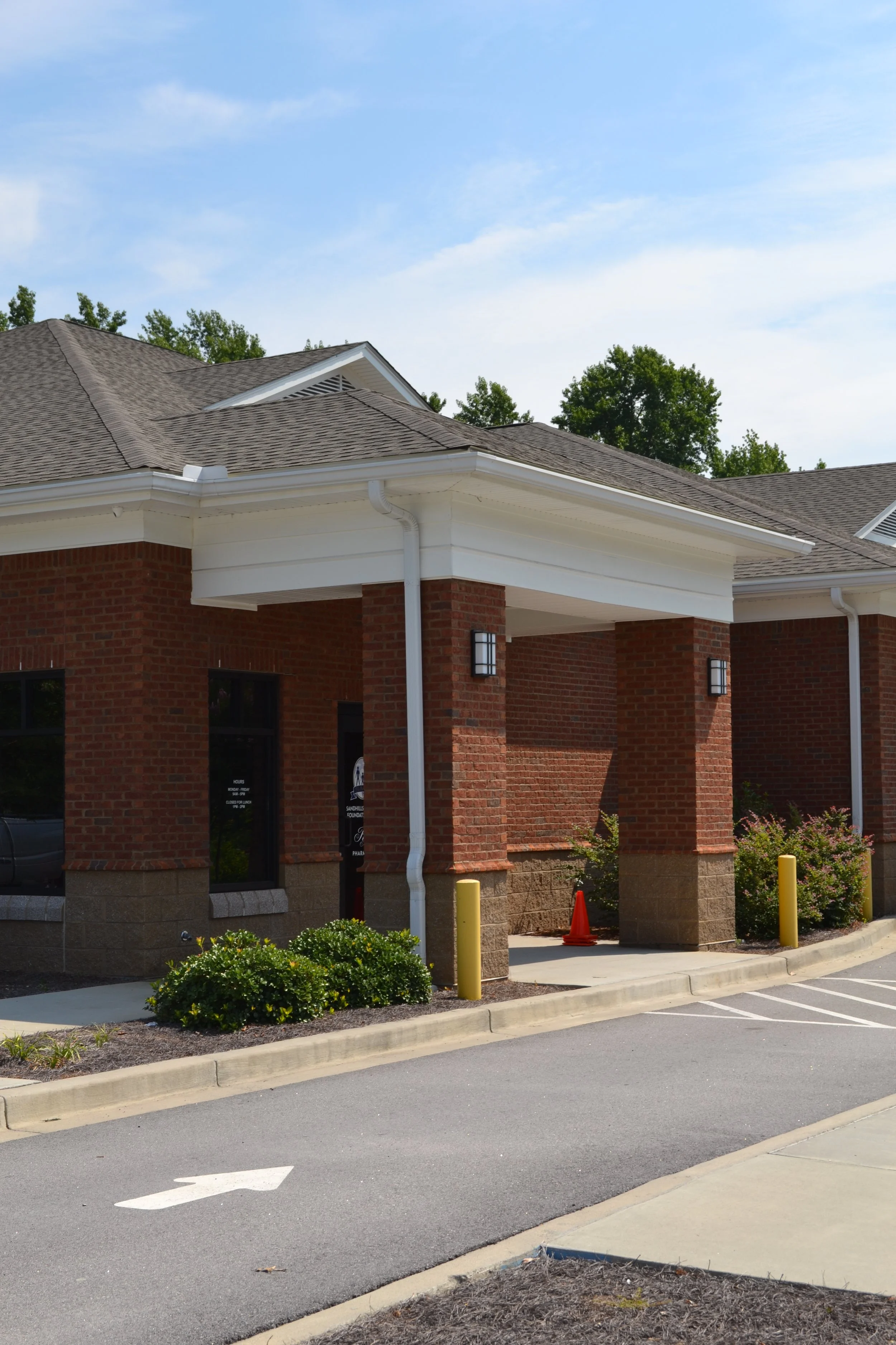 View of a brick building with white trim and multiple roofs, part of a parking lot with yellow posts and a white arrow painted on the asphalt, green bushes and trees in the background under a blue sky.