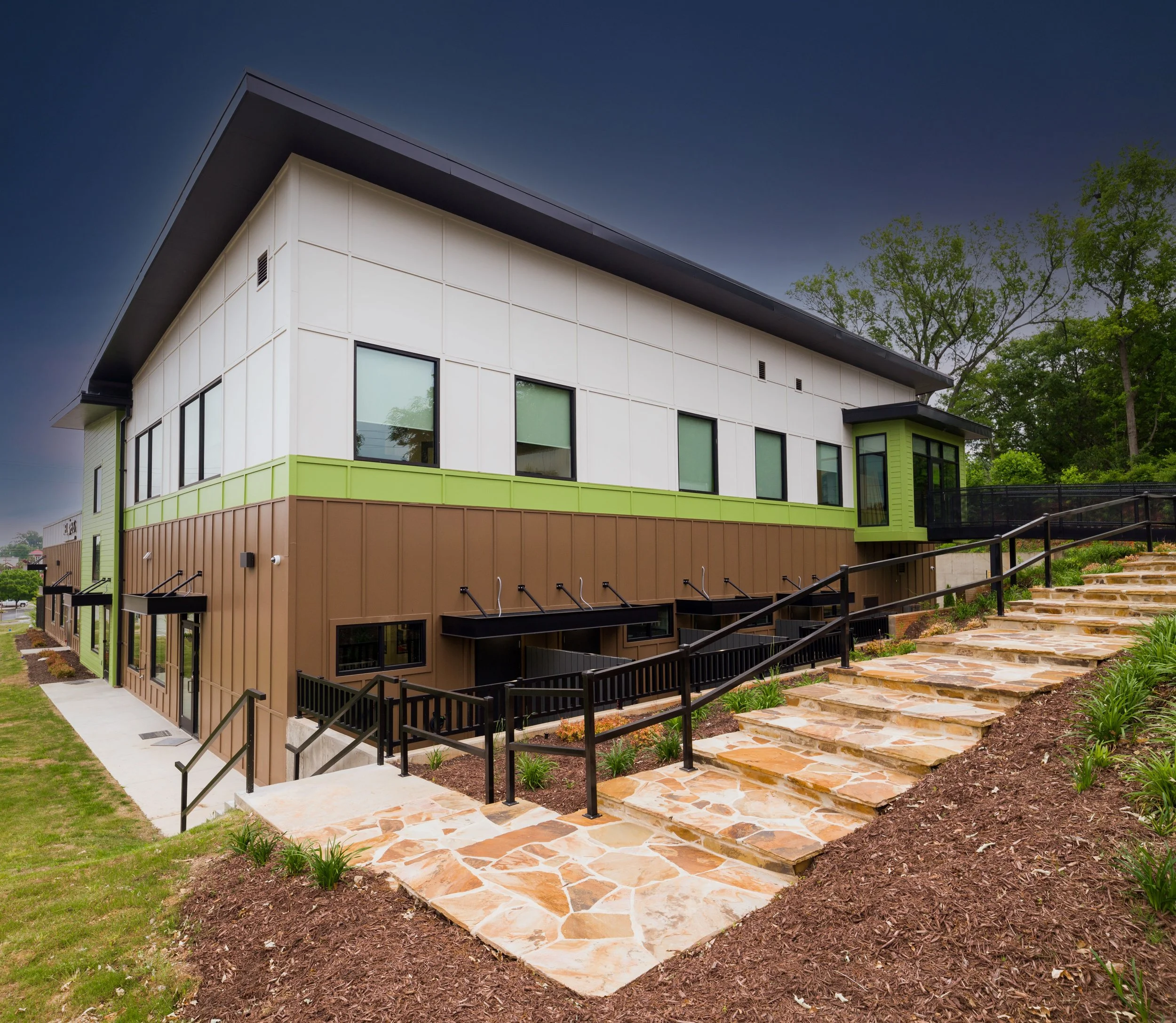 Modern building with large windows, tan and green exterior, stone pathway, black railings, surrounded by landscaping and trees, under a dark sky.