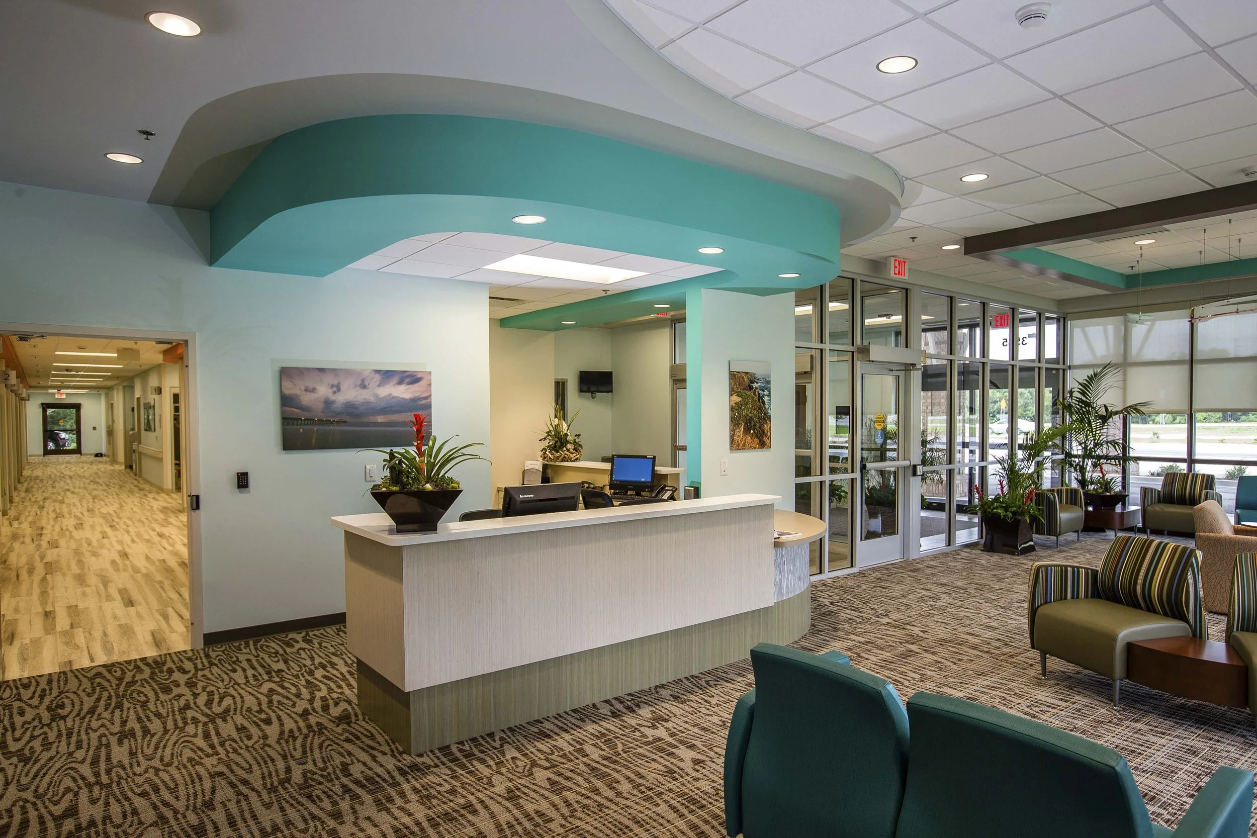 A modern hospital or medical facility lobby with a front desk, seating area, glass doors, and green plants.