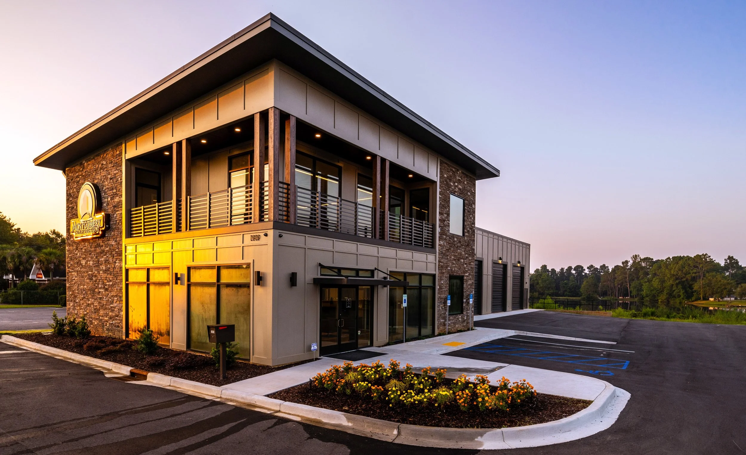 A two-story commercial building with a brick and beige exterior, a balcony with a metal railing on the second floor, and a sign that reads 'Montgomery Construction'. The parking lot in front has several marked parking spaces and some flowerbeds with yellow and orange flowers, with a clear sky at dusk.