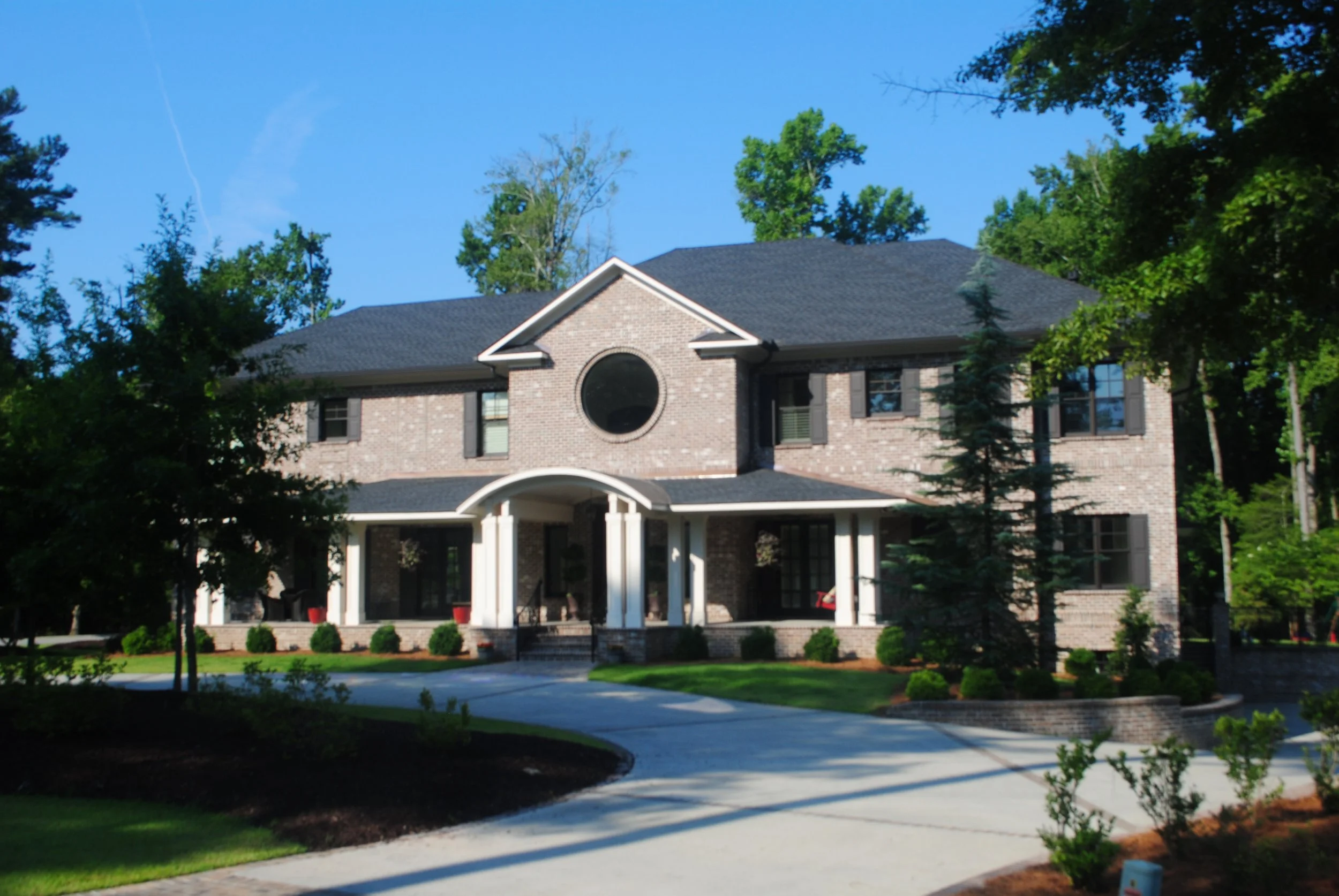 A large brick house with a dark gray roof, white columns at the entrance, and black shutters on the windows. The house is surrounded by green trees, bushes, and a well-maintained lawn with a curved concrete walkway leading up to the front door. The s