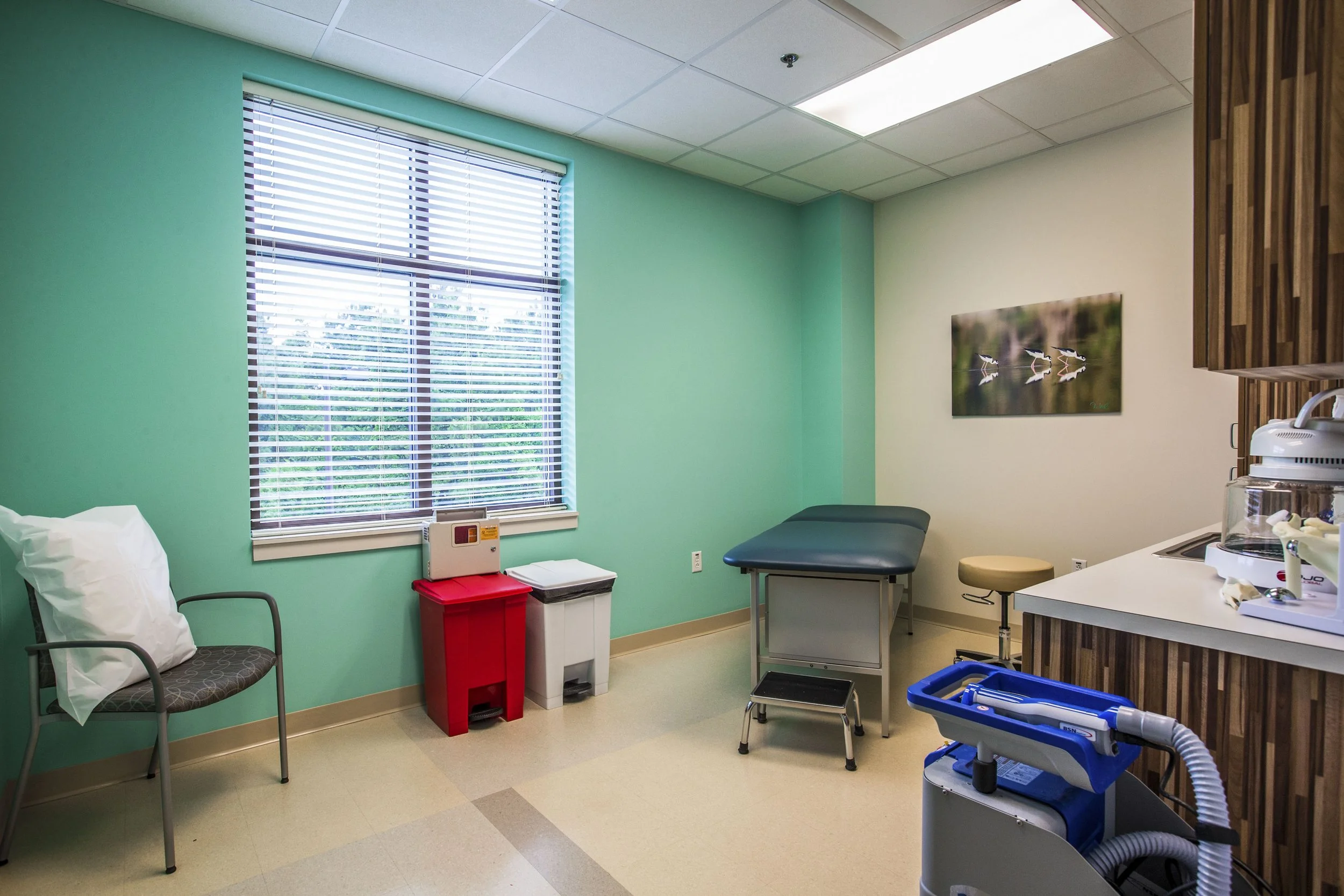 A medical examination room with a window, teal wall, examination table, medical equipment, and chairs.