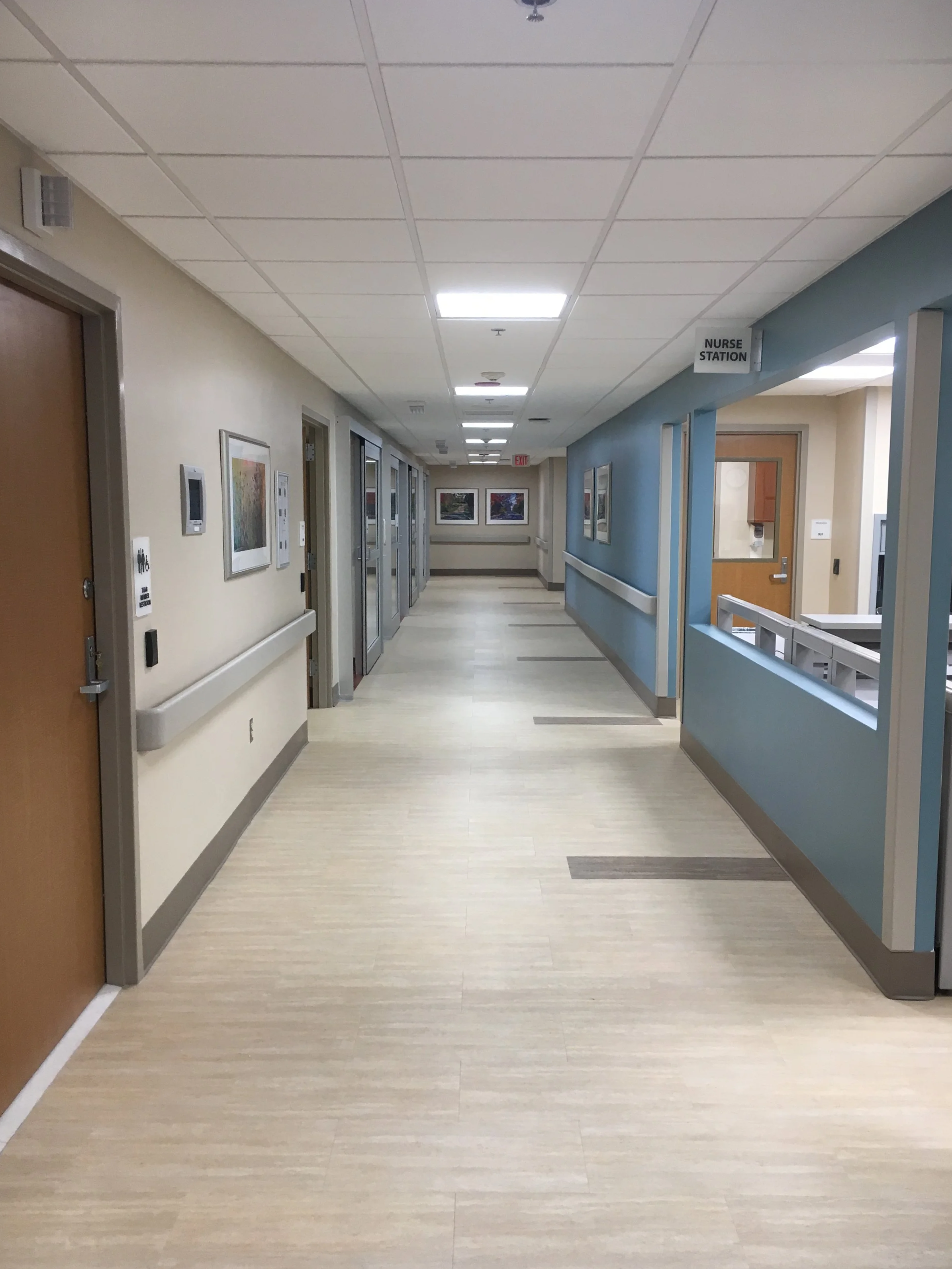 Hospital hallway with beige walls, framed artwork, and a blue wall with a sign that says 'NURSE STATION'. Wooden doors, elevator doors, and a windowed door insert leading to nurse's station visible.