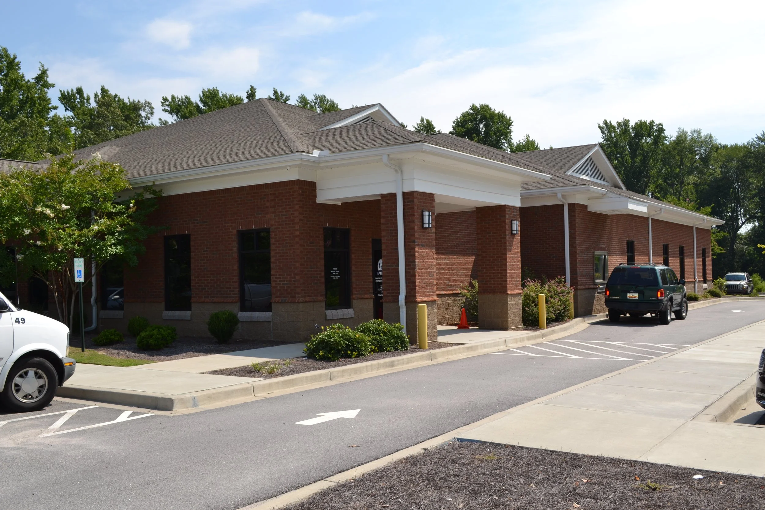 A brick commercial building with white trim and a sloped brown shingle roof, surrounded by green trees and parked cars, in a parking lot with marked spaces.