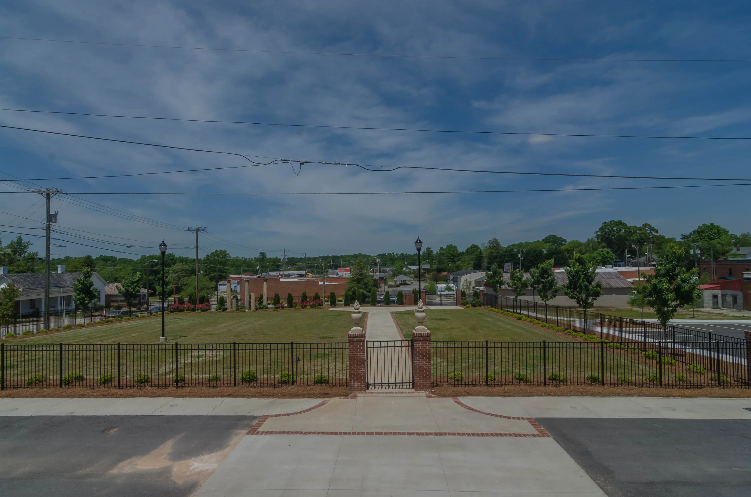 A large, well-maintained green lawn with a walkway leading to a monument with statues, surrounded by a black iron fence. In the background, there are houses, trees, and a row of utility poles with power lines, under a partly cloudy sky.