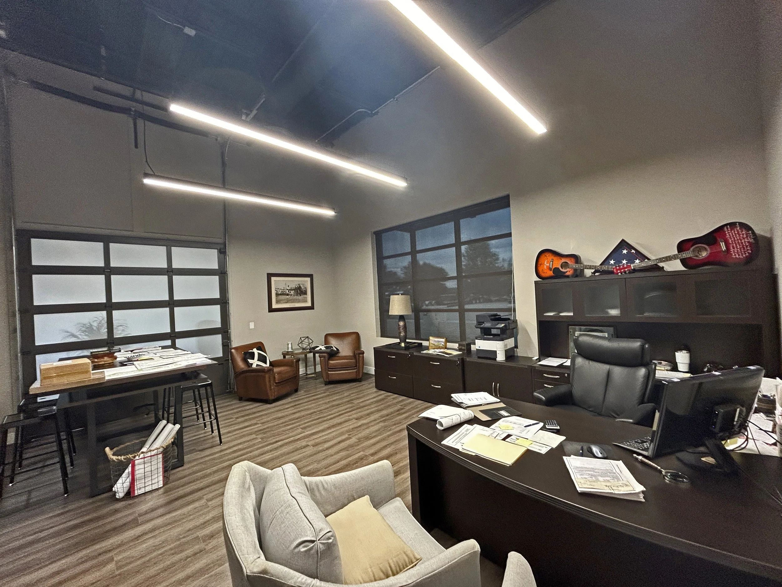 An office room with a large window, a desk cluttered with papers, a computer, a black office chair, a beige armchair with a pillow, two brown chairs, a cabinet with guitars on top, and fluorescent ceiling lights.