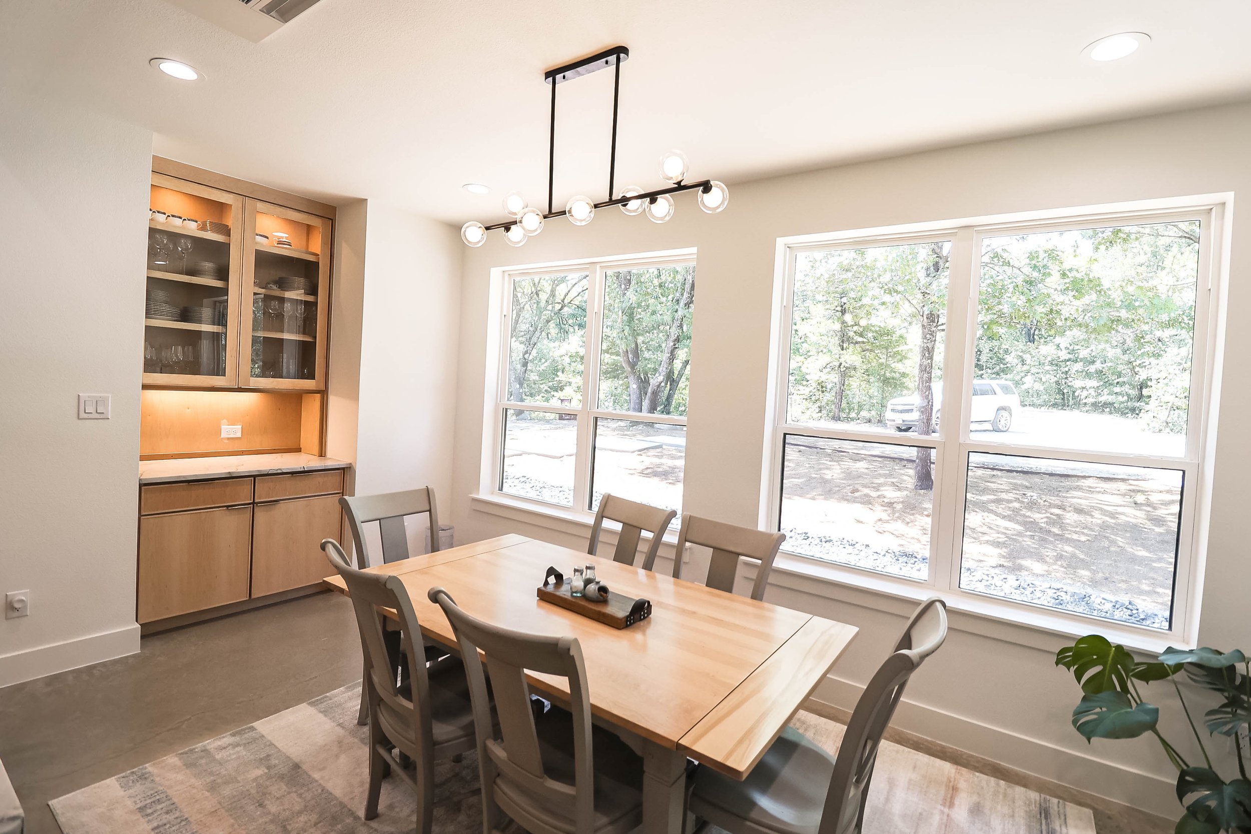A dining room with large windows showing trees outside. There is a rectangular wooden table surrounded by eight light gray chairs. A black and glass chandelier hangs above the table. To the left, an open cabinet displays dishes and glassware. The roo