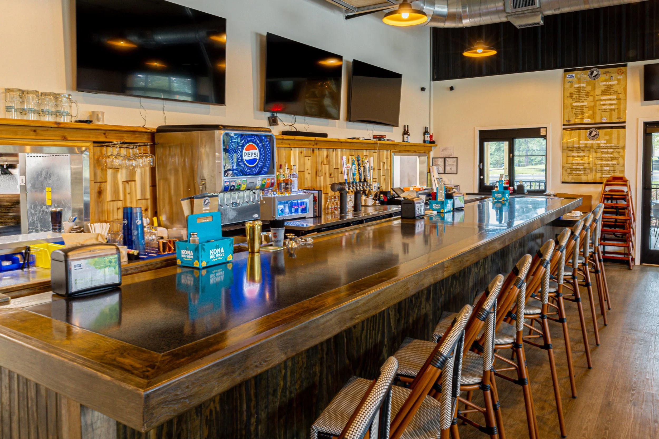 Interior of a restaurant or bar with a long wooden counter, bar stools with woven seats, and multiple flat-screen TVs on the wall. There is a Pepsi soda fountain, liquor bottles, and glassware on the counter. The space features industrial lighting and large windows.
