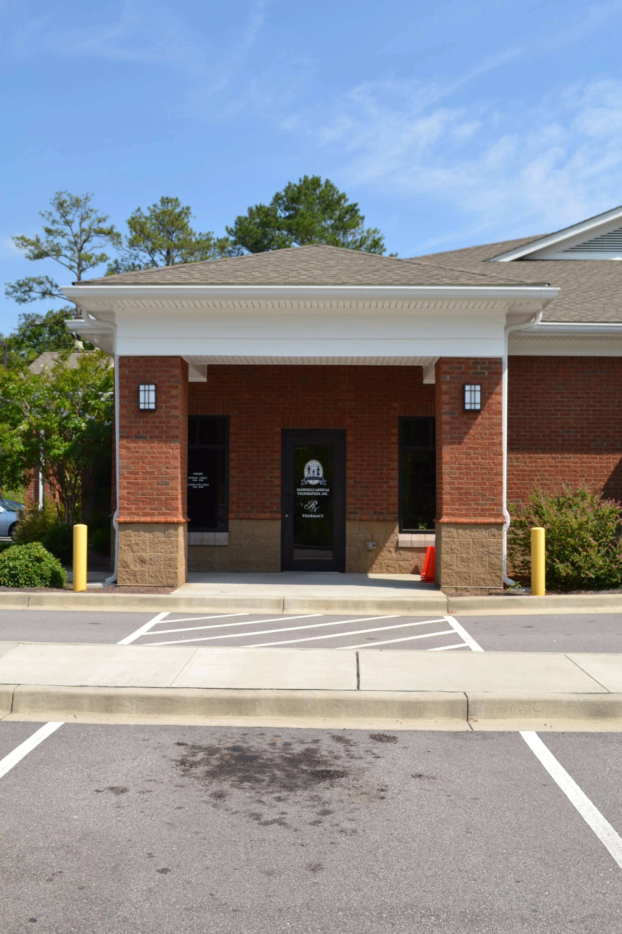 Front view of a brick building with a covered entrance and glass door with logo, flanked by two yellow posts, parked cars and a landscaped area in front, under a blue sky with trees in the background.