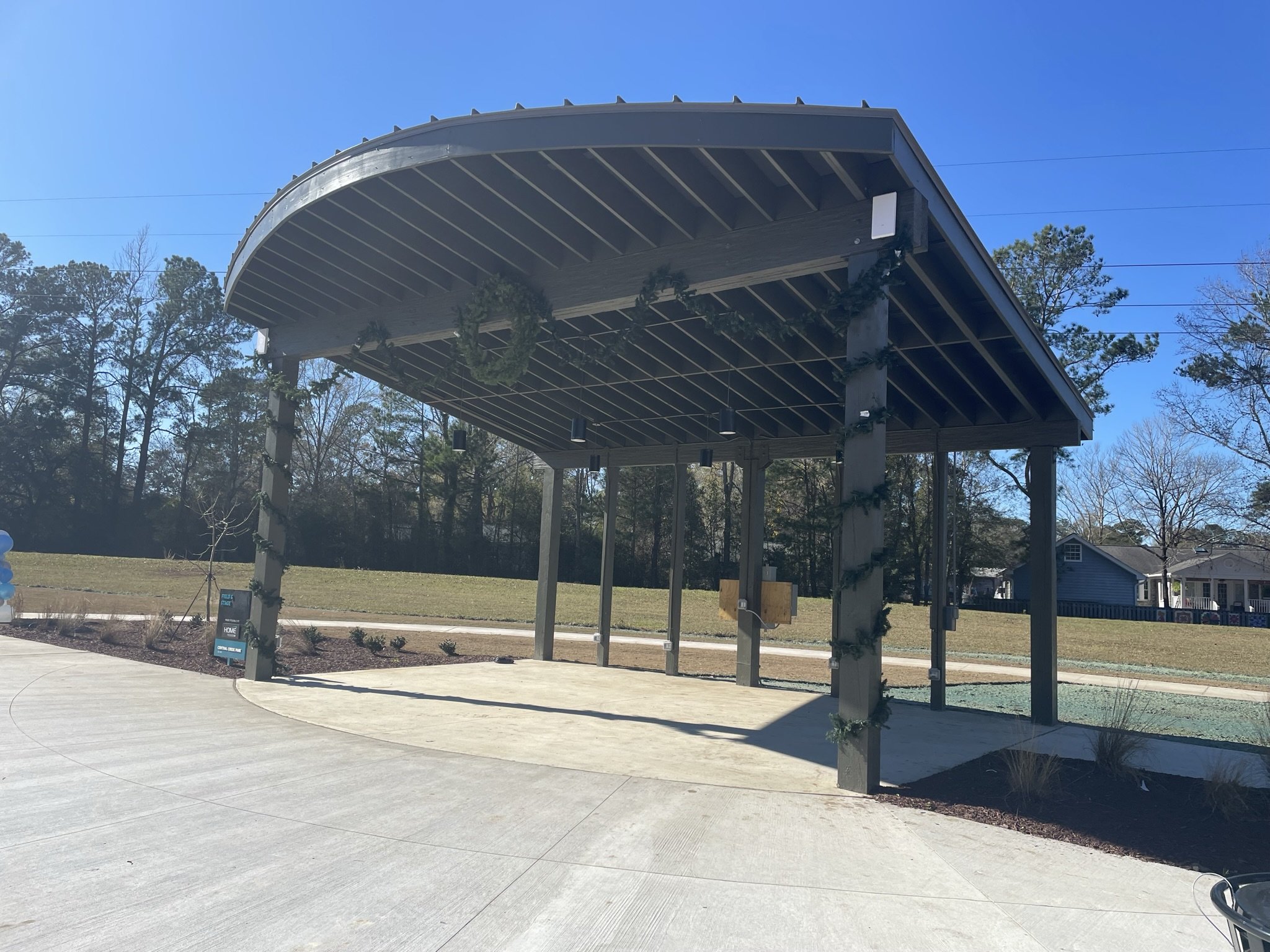 A modern outdoor stage with a curved roof decorated with holiday garland, set in a park with trees and houses in the background under a clear blue sky.