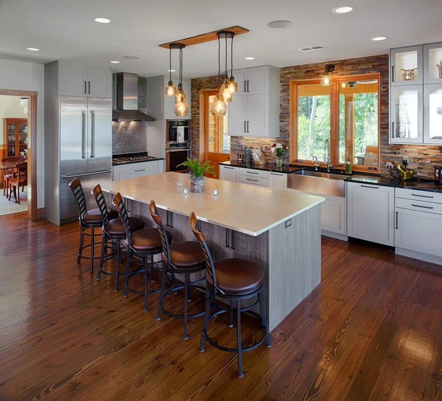 Modern kitchen with white cabinets, a large island with a light-colored countertop, four brown leather bar stools, a brick accent wall, and a window overlooking greenery.