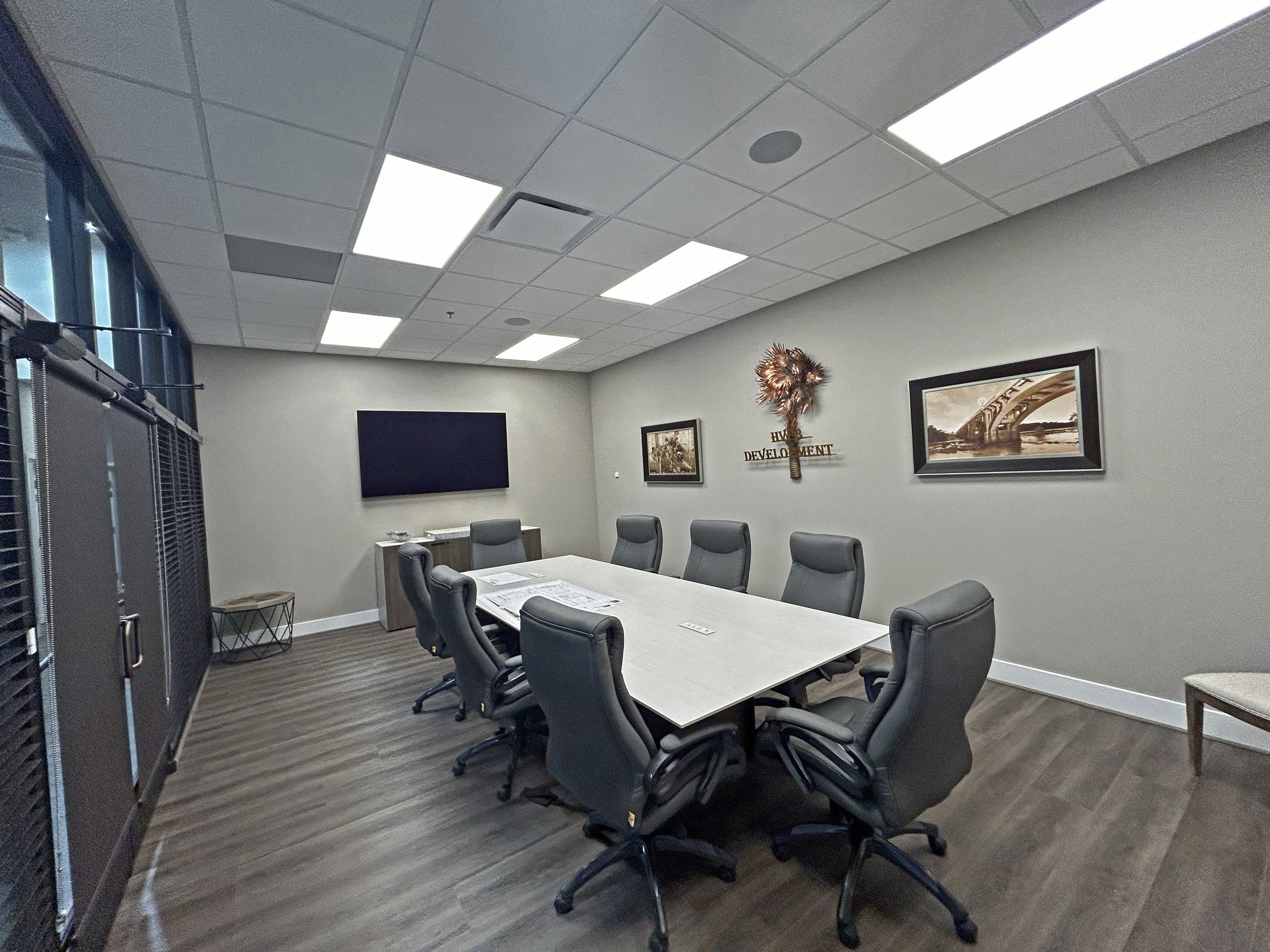 A conference room with a large rectangular table surrounded by black office chairs. The room has a ceiling with recessed fluorescent lighting, a flat screen TV on the wall, and framed pictures. There are windows covered with blinds on one side, and t