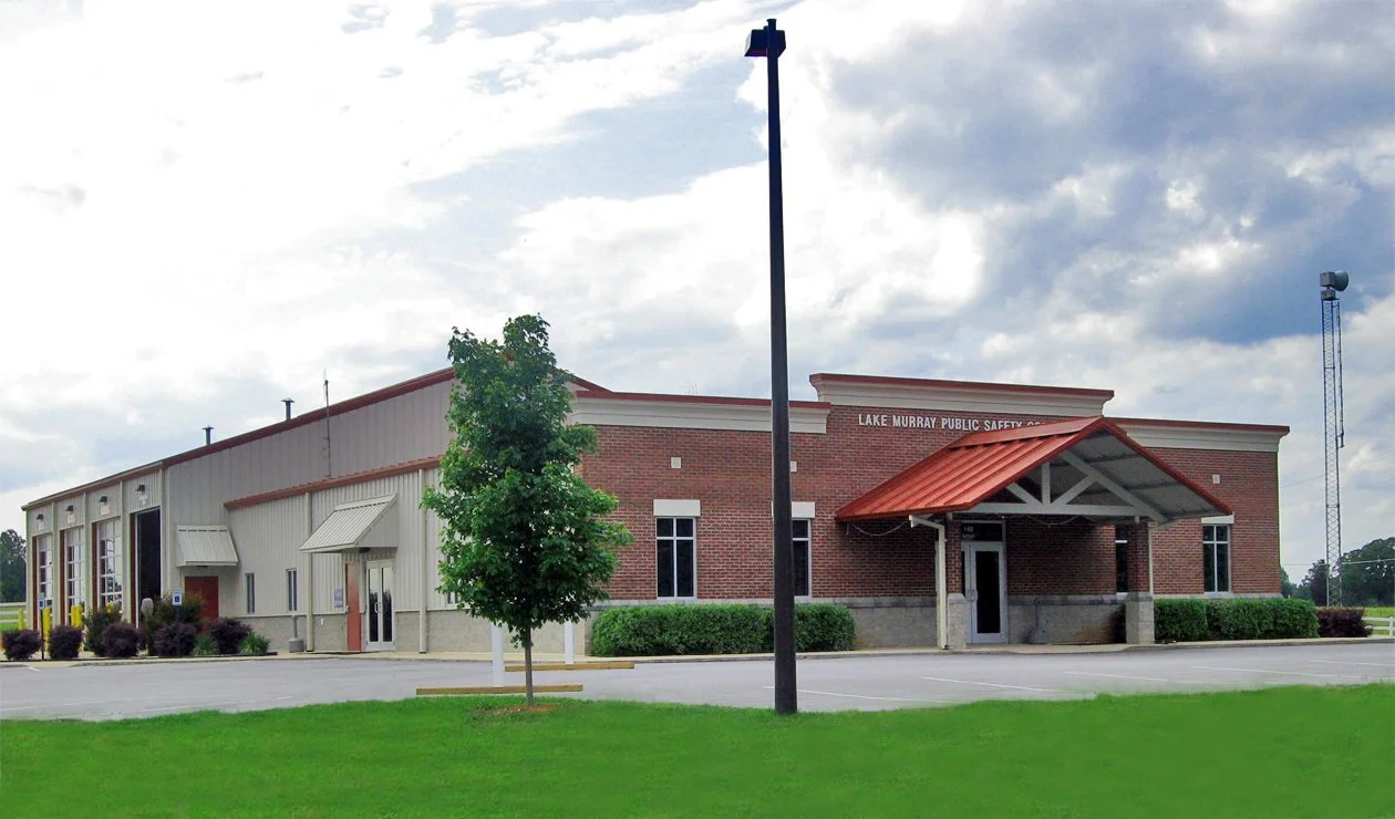 Public safety building with a brick and metal facade, labeled Lake Murray Public Safety, with a small parking lot and a landscaped area with a small tree and grass.