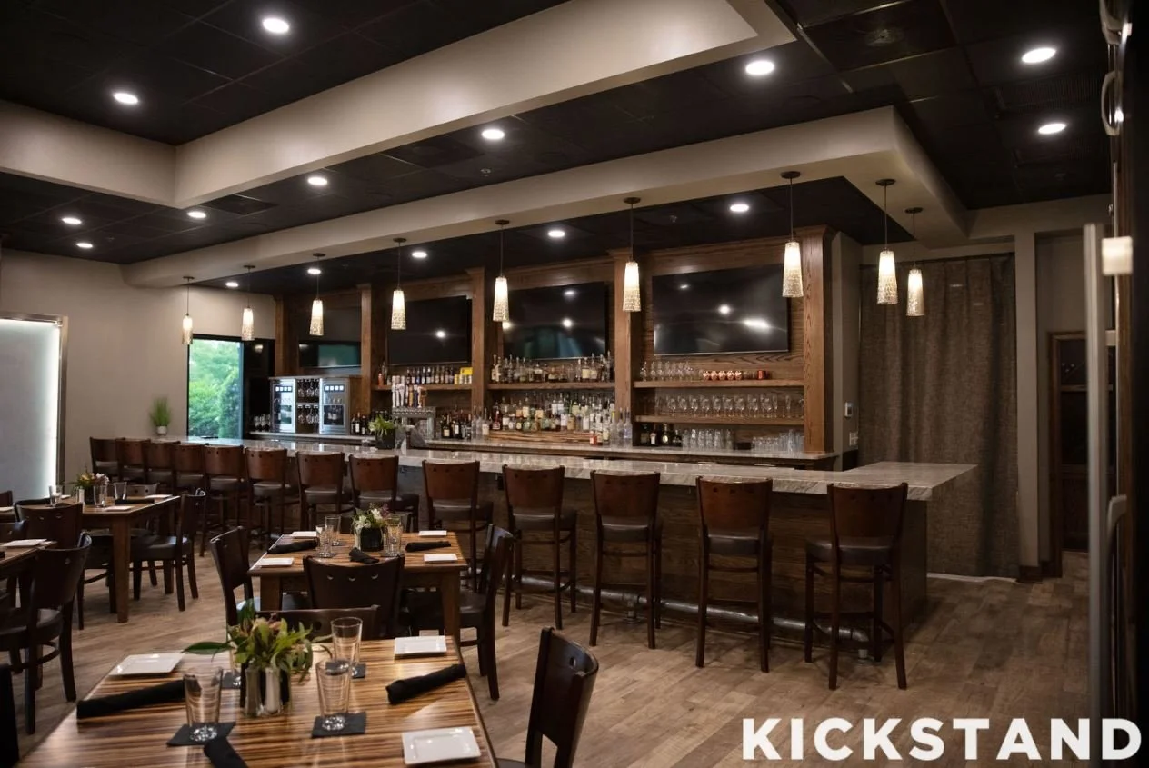 Elegant restaurant with a bar area featuring bar stools, wine glasses, liquor bottles, and two large televisions on a wood-paneled wall, with dining tables set with napkins, glasses, and flower centerpieces.
