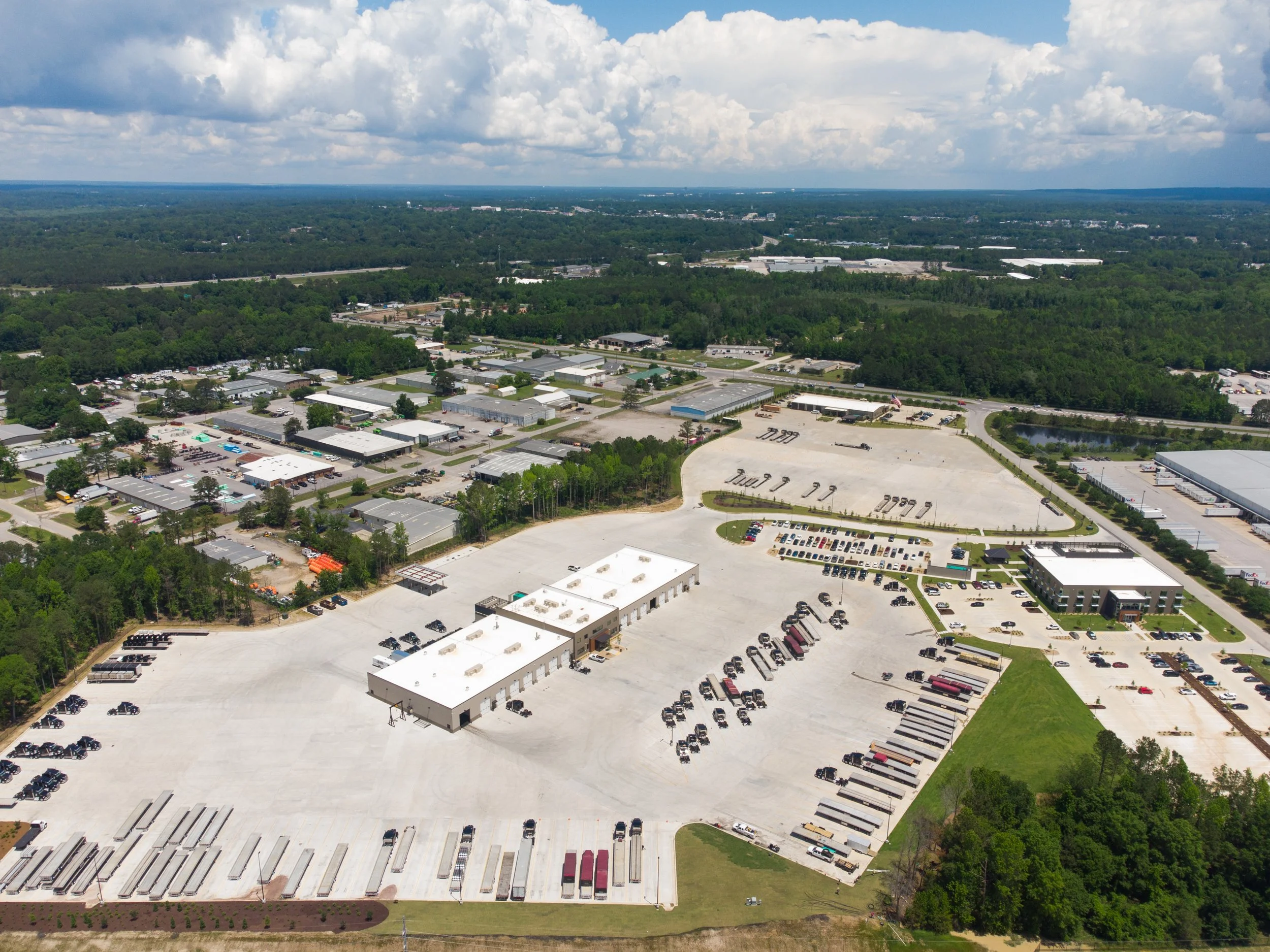 An aerial view of a commercial and industrial area with numerous buildings, parking lots, and a green wooded area in the background under a partly cloudy sky.
