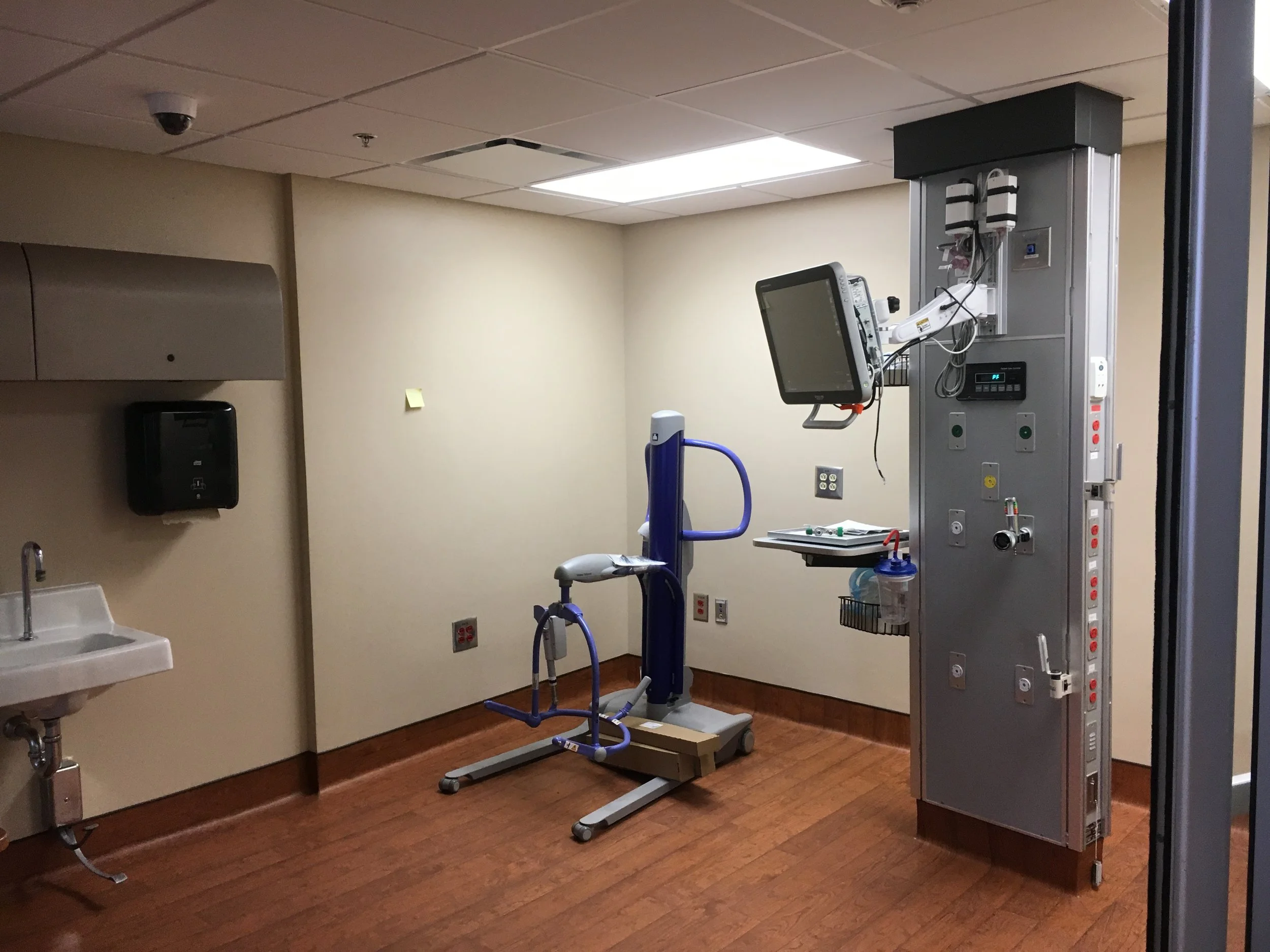 Hospital room with medical equipment including a patient monitor, IV stands, and a standing lift device, with wood flooring and beige walls.