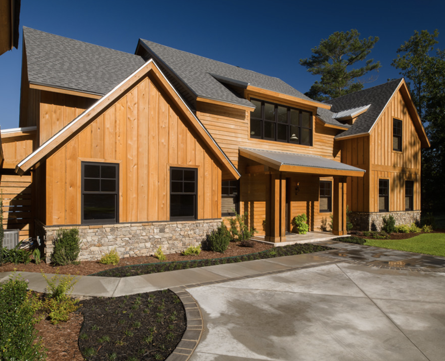 Modern two-story house with wooden siding, stone foundation, gray roof, and black-framed windows, surrounded by landscaping and a concrete driveway.