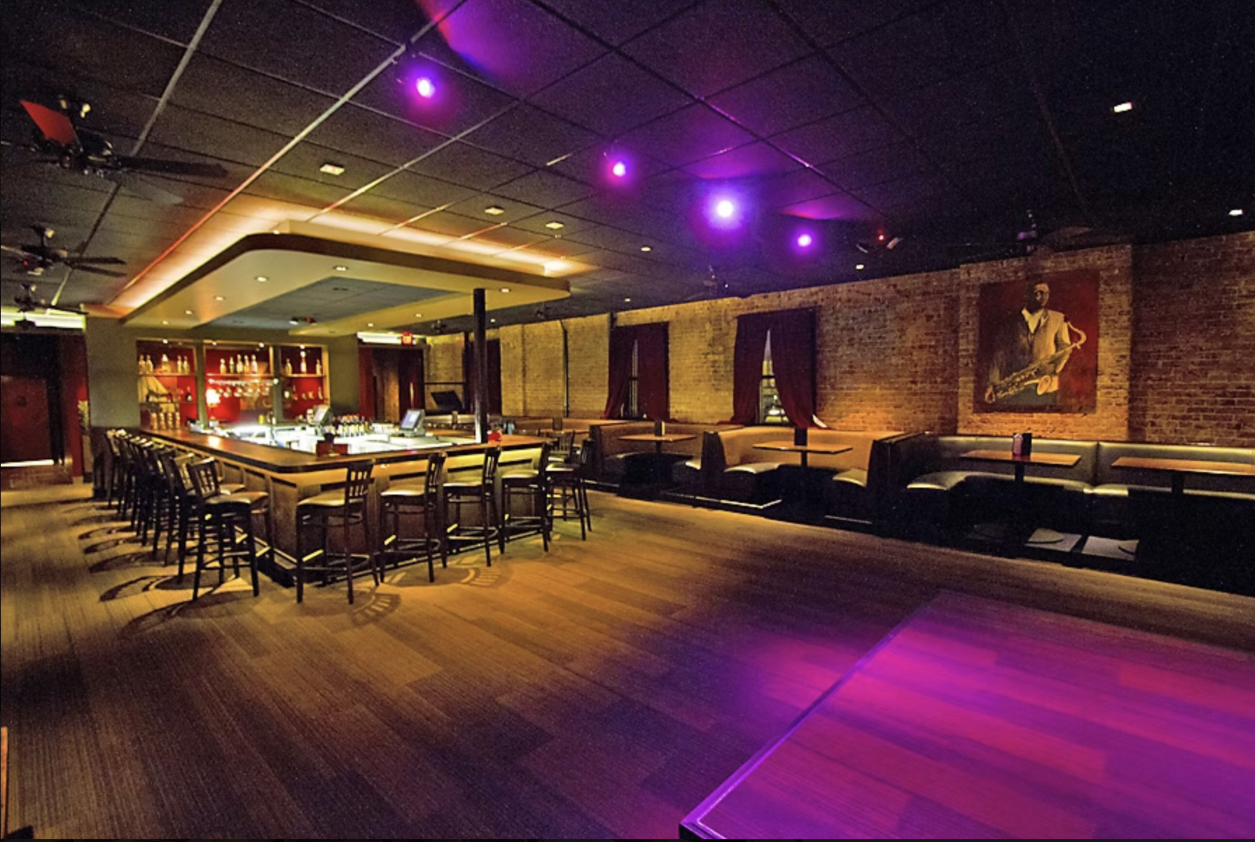 Interior view of a dimly lit bar and lounge with a rectangular bar counter, bar stools, red shelving with bottles, brick walls, and a jazz-themed painting on the wall. Colorful purple and pink ceiling lights illuminate the space.
